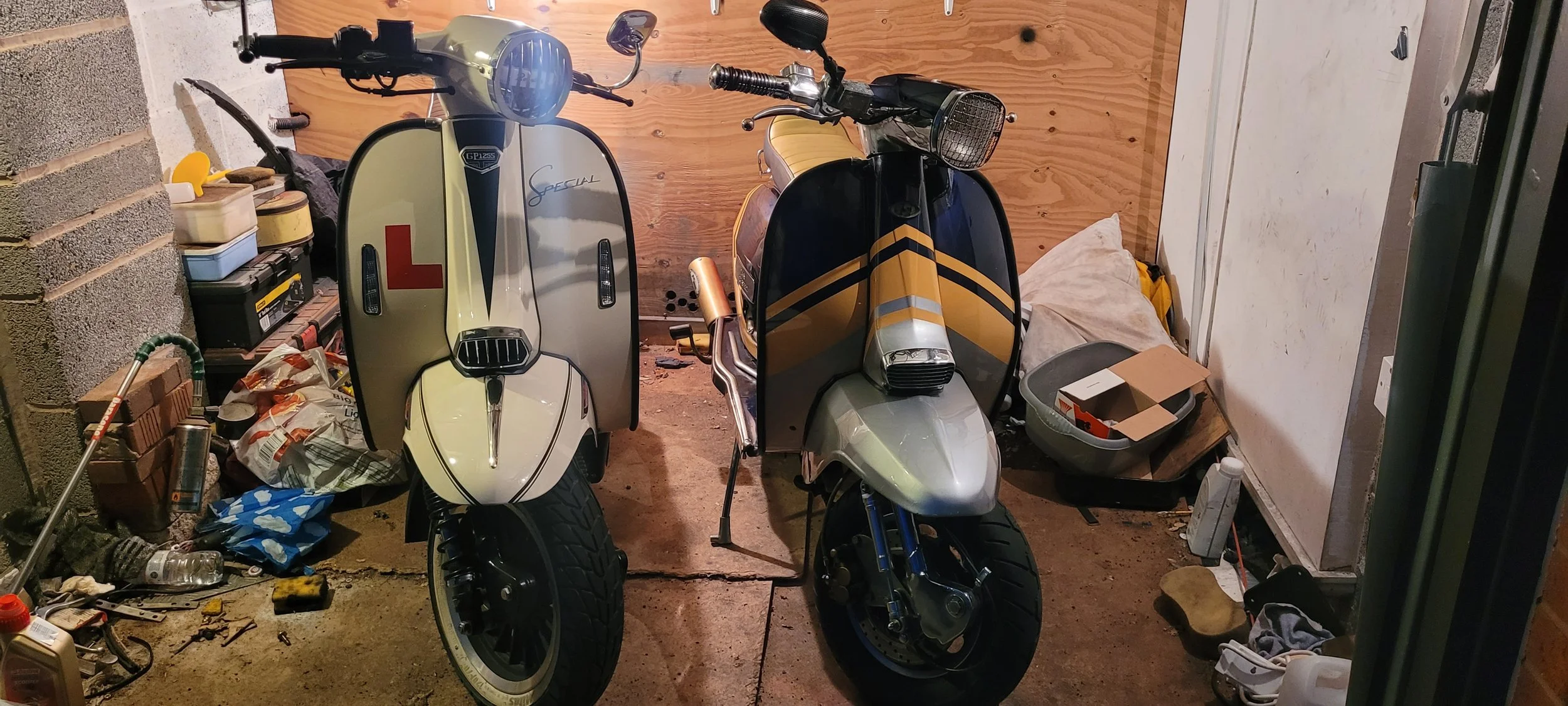 Two vintage scooters parked side by side in a garage, surrounded by various tools, storage boxes, and miscellaneous items.