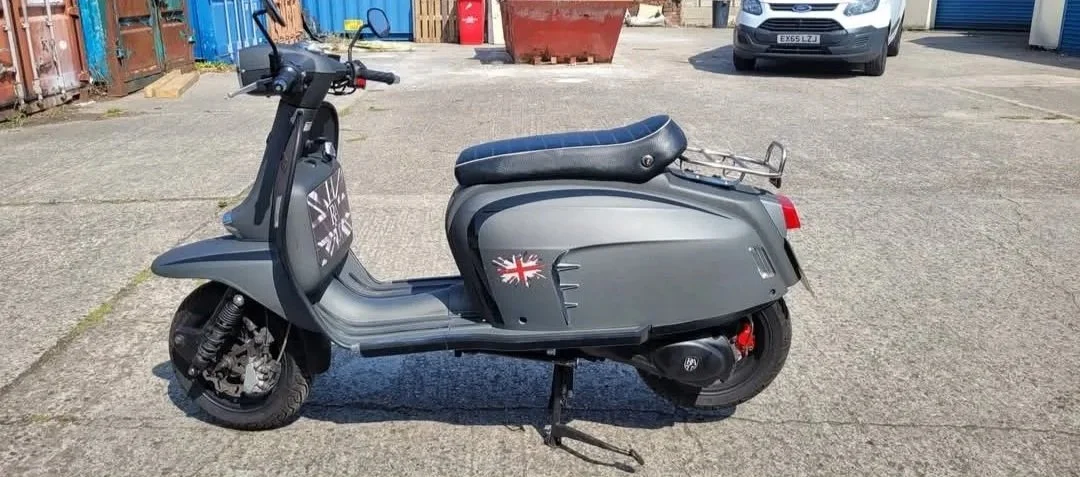 Gray vintage scooter with a seat, parked on a concrete surface in an outdoor lot, with other vehicles and building structures in the background.