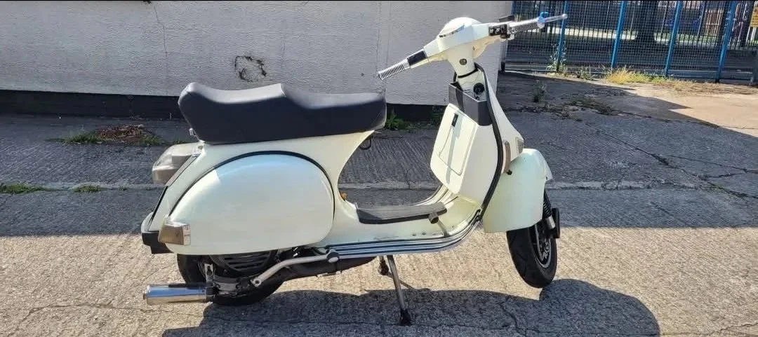 White vintage scooter parked on a sidewalk near a wall and a fence.
