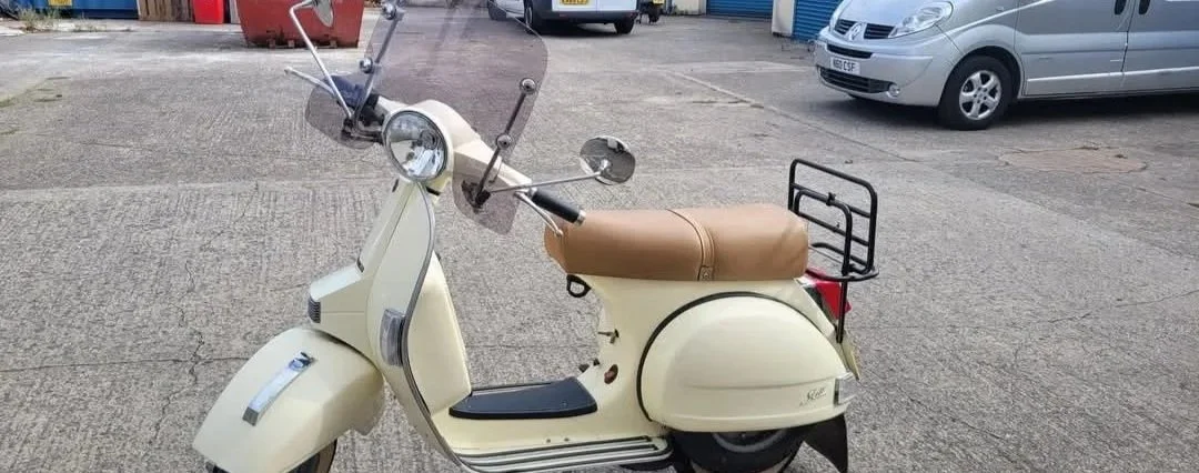 Cream-colored vintage scooter with a tan seat and a small rear cargo rack, parked on a concrete lot with cars in the background.