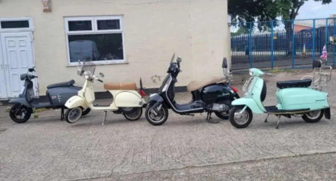 Four vintage scooters parked in a row on a sidewalk in front of a beige building with a window and door, and a blue fence in the background.