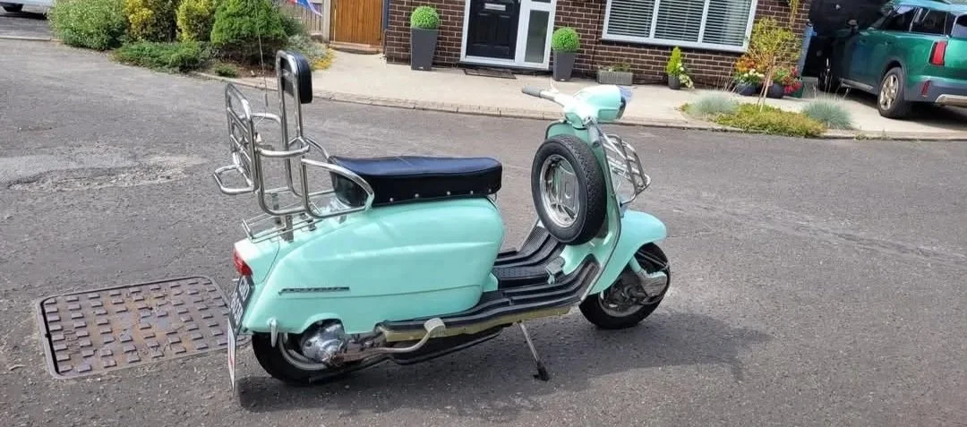 A vintage mint green scooter with a small spare tire mounted on the front, parked on a residential street.