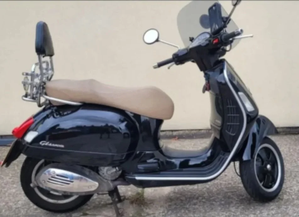 Black scooter with a beige seat and a backrest, parked against a plain wall.