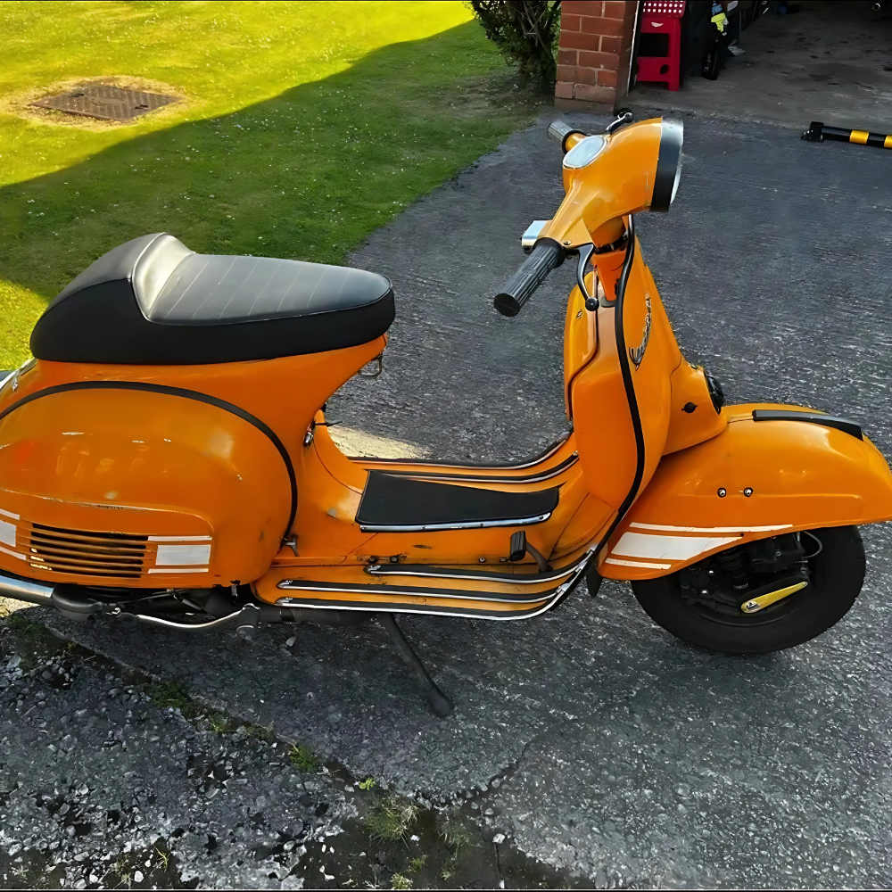 Orange vintage scooter parked on a gravel driveway next to a grassy lawn and a brick garage entrance.