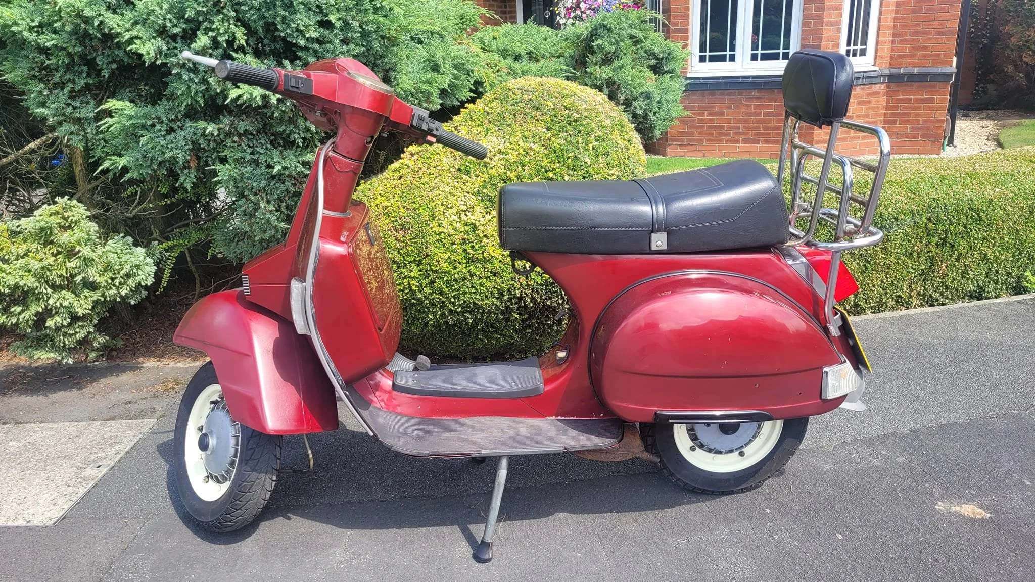 A vintage red scooter parked on a driveway with green bushes and a brick house in the background.