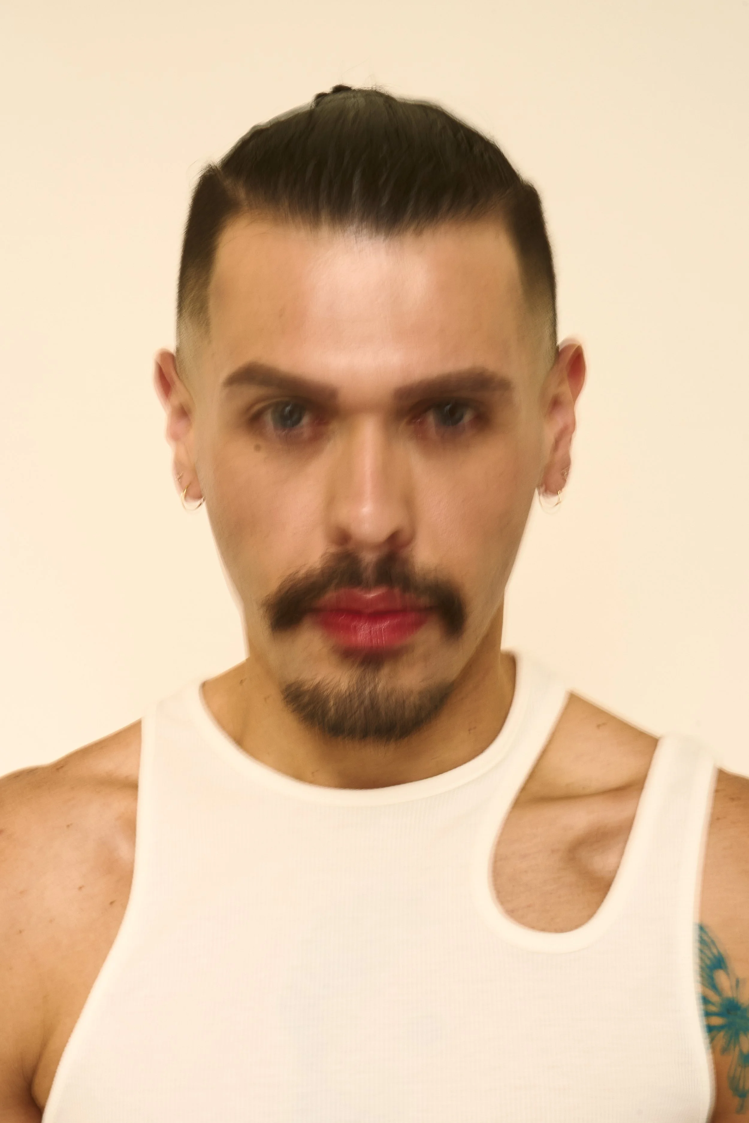 A person with short dark hair, a mustache and goatee, wearing earrings, and a white sleeveless top, facing forward against a light background.