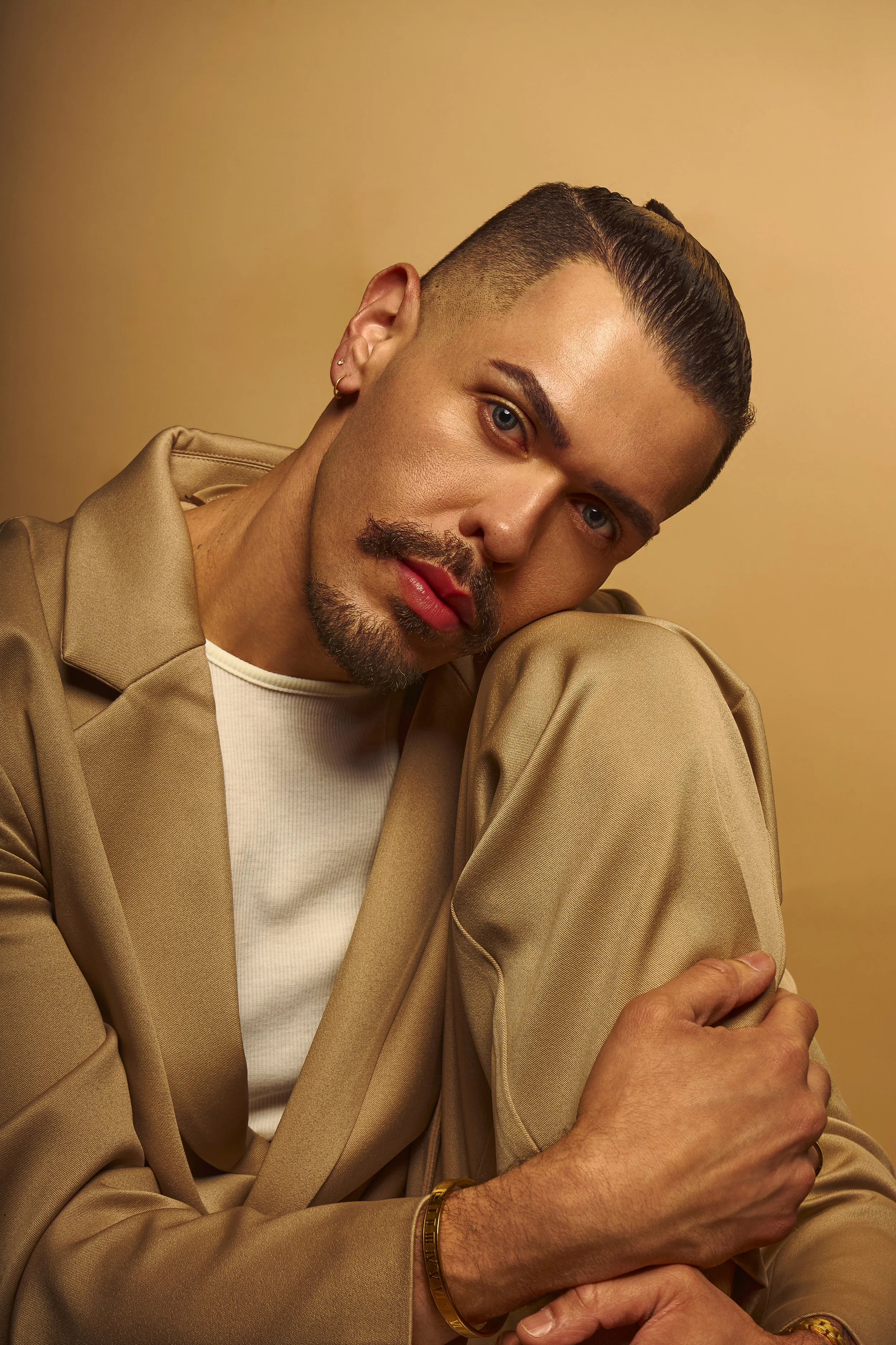 A man with light skin, slicked-back hair, and a goatee, wearing a beige blazer and a white shirt, resting his head on his knee against a beige background.