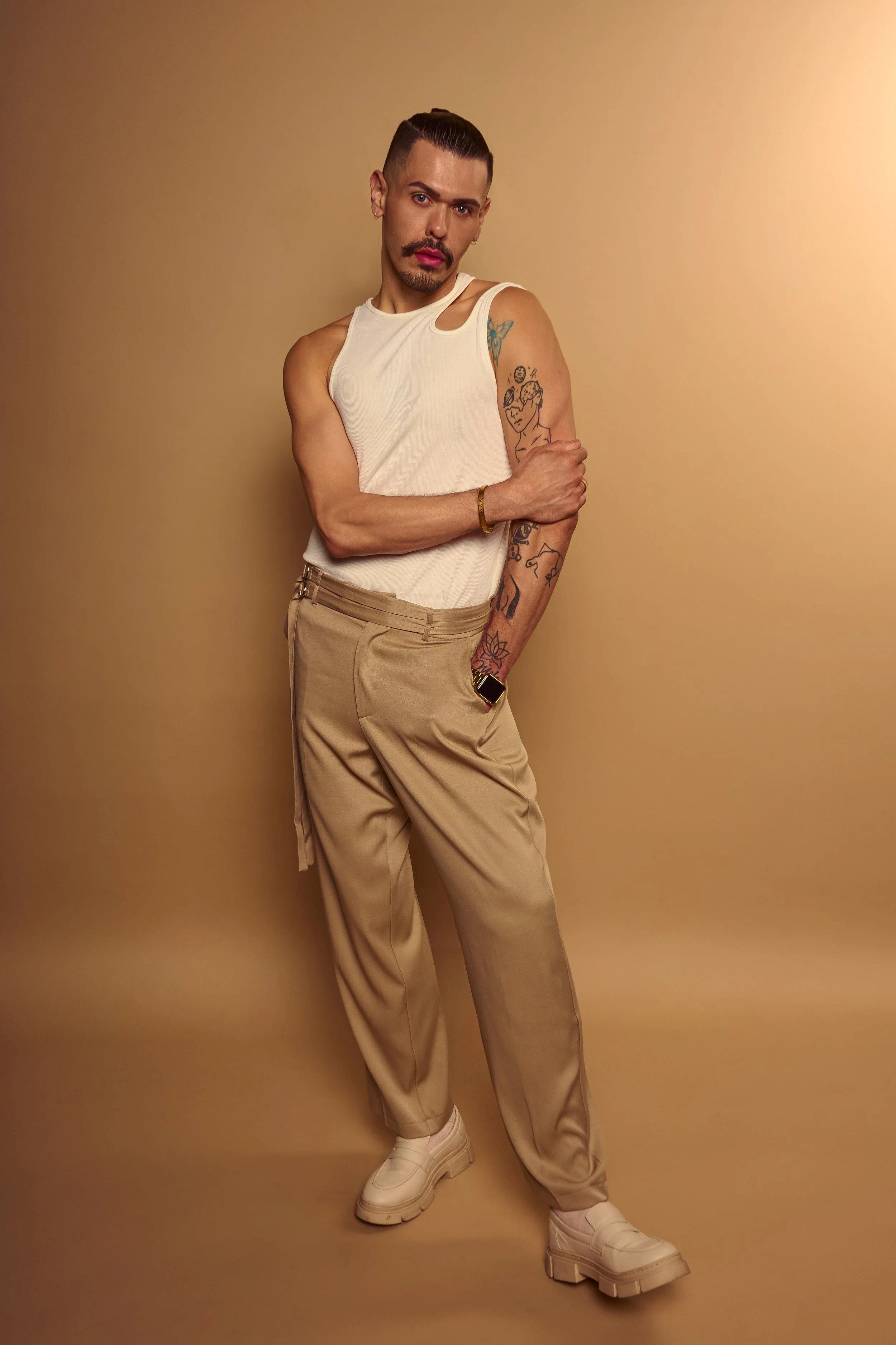 A person standing against a beige background, wearing a sleeveless white top, beige pants, and chunky white shoes. They have tattoos on their right arm, a mustache, and a goatee, with slicked-back hair.