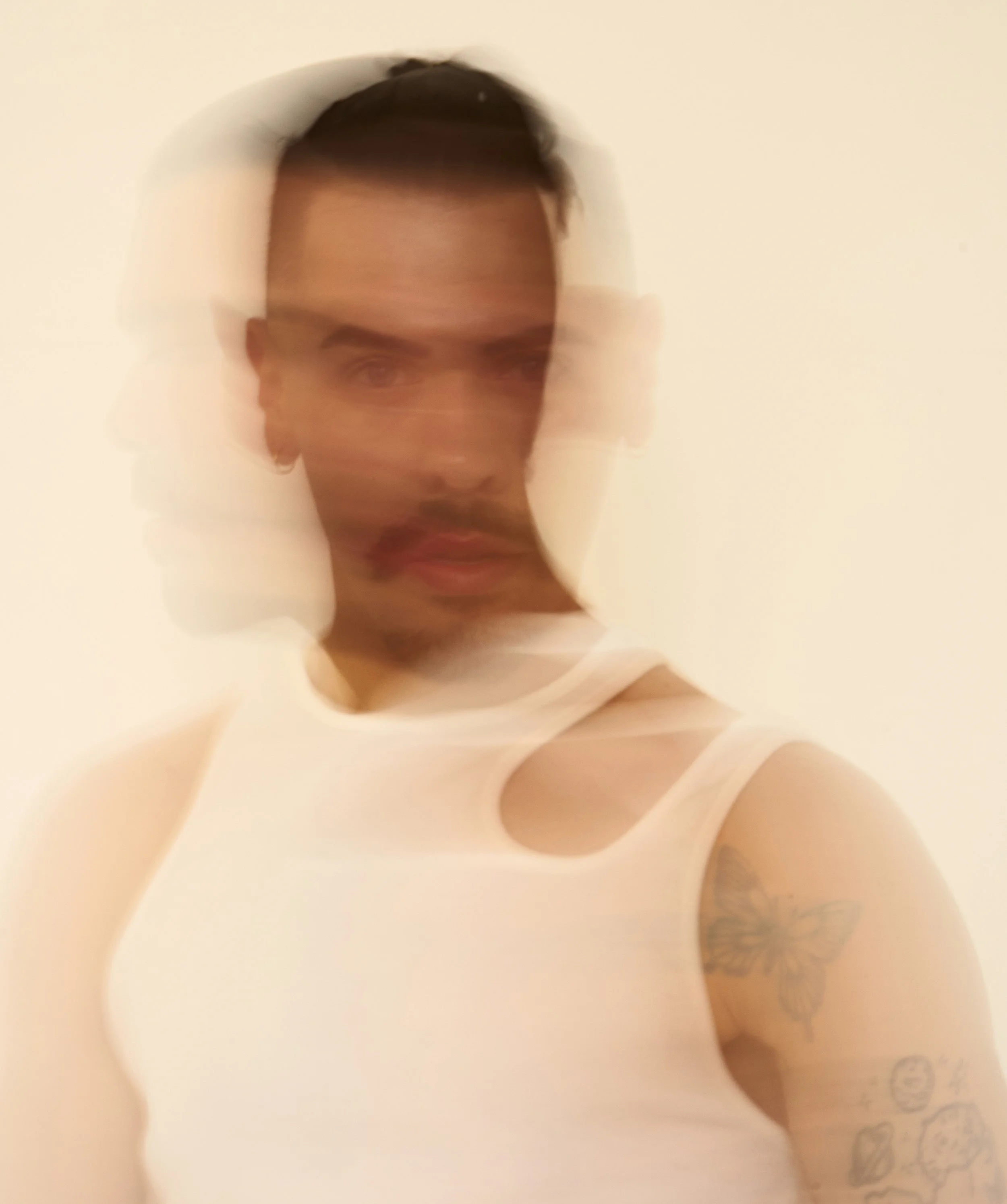 A person with short dark hair, a mustache, and a goatee, wearing a sleeveless white top, with tattoos on their arm, taking a blurry selfie against a plain white background.