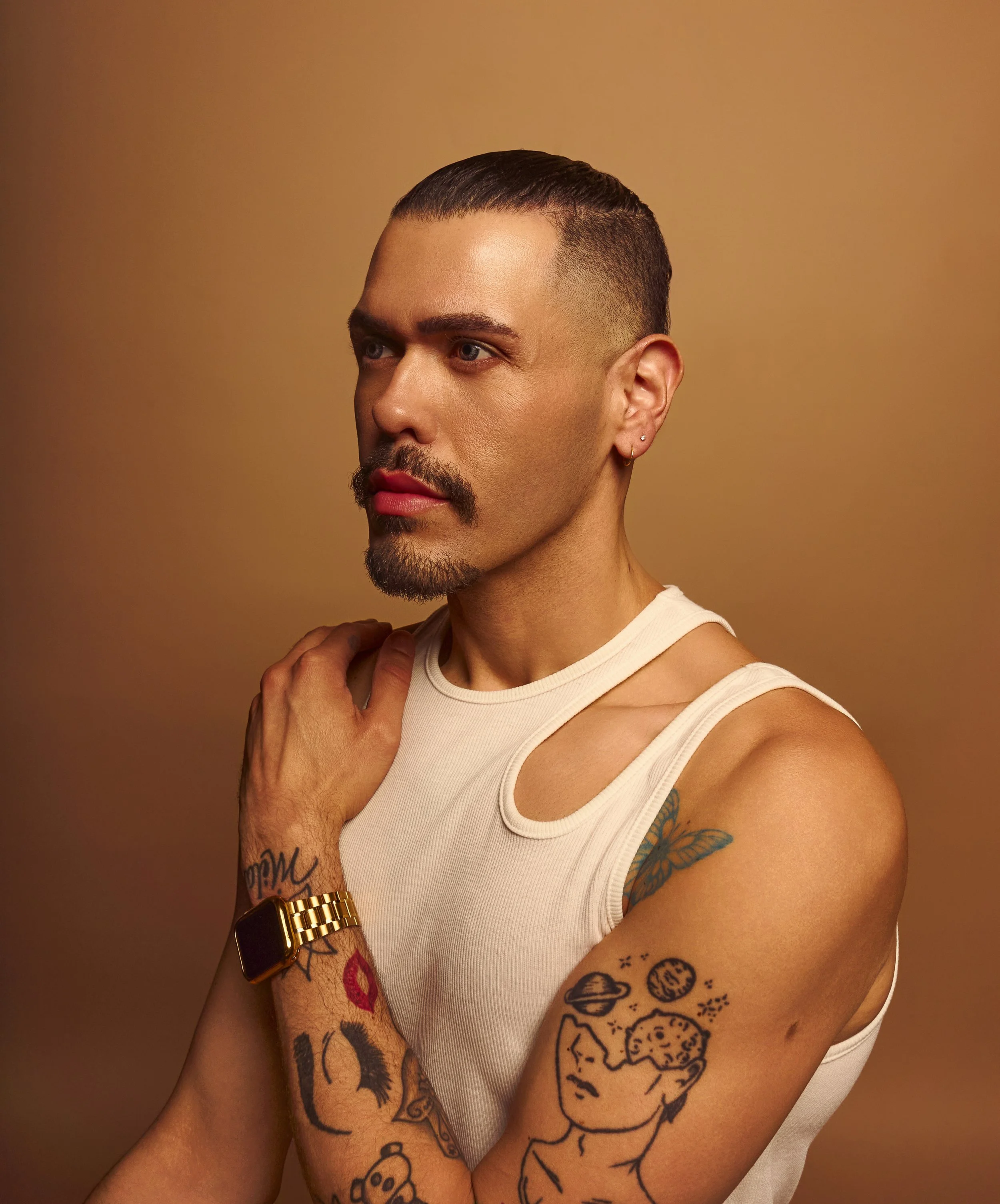 A man with a beard and mustache wearing a white sleeveless shirt, gold watch, and multiple tattoos, looking to the side with a neutral expression, against a brown gradient background.
