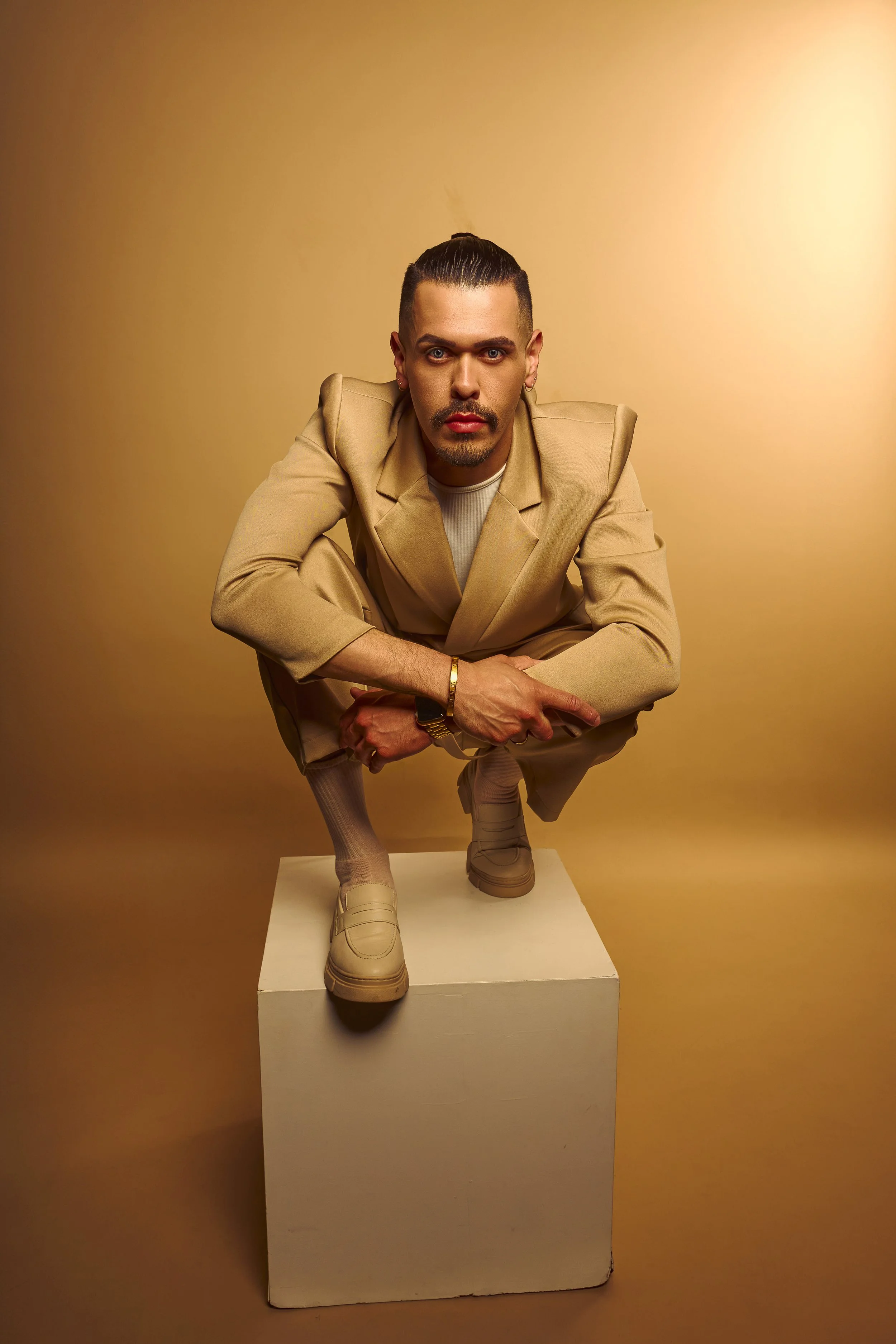 A man squatting on a white block against a beige background, wearing a tan suit, beige sneakers, and accessories, looking directly at the camera with intense expression.