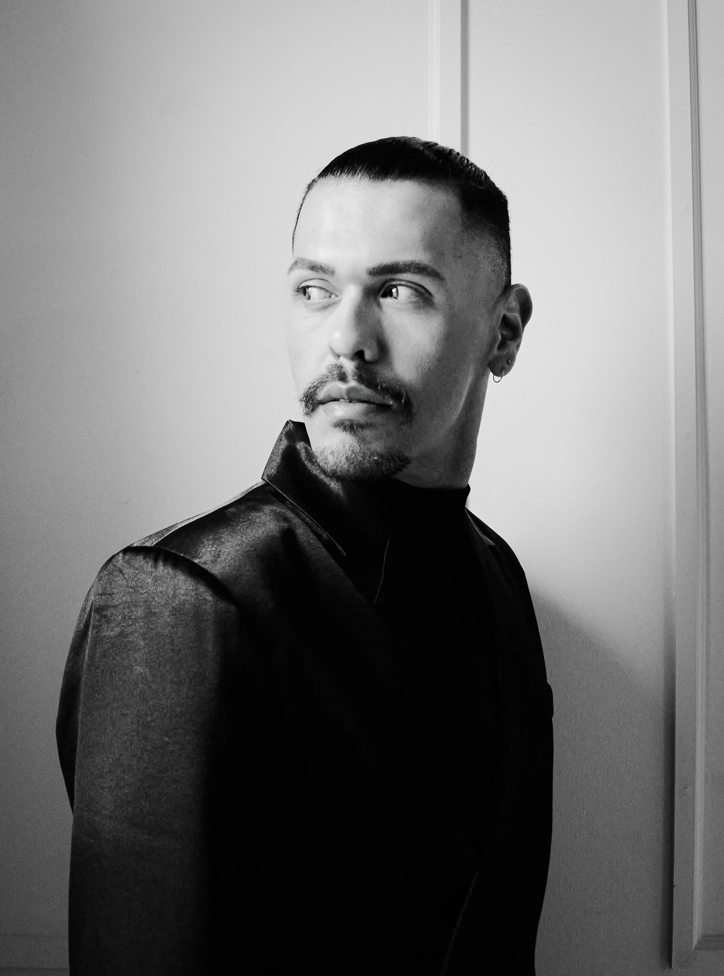 A black and white portrait of a man with a slicked-back hairstyle and facial hair, looking to the side, wearing a dark shirt, standing in front of a plain background.