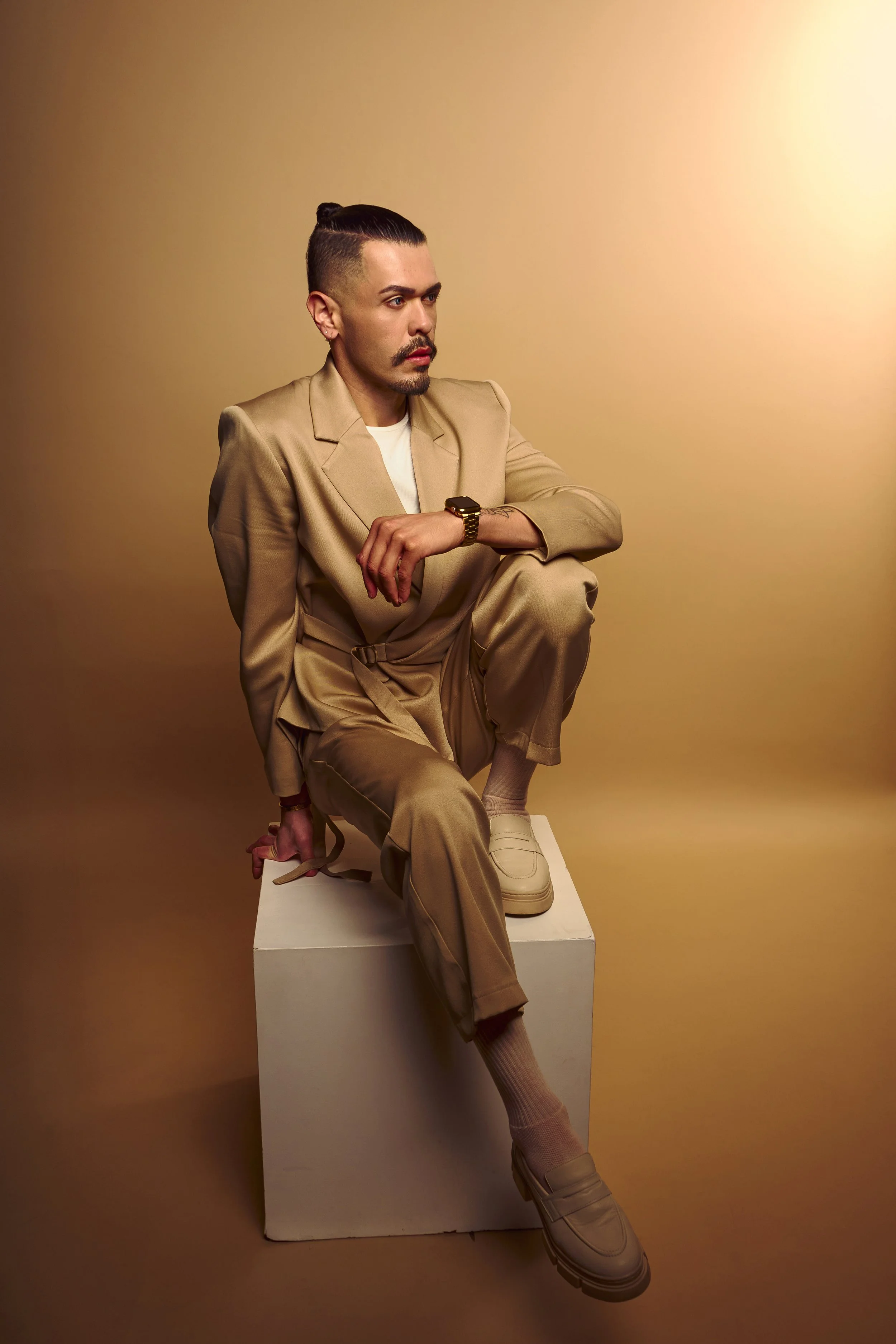 A man with a mustache and beard, wearing a beige suit, tan shirt, and beige shoes, sits on a white block against a yellowish background. He is wearing a watch on his left wrist and has his hair tied back.