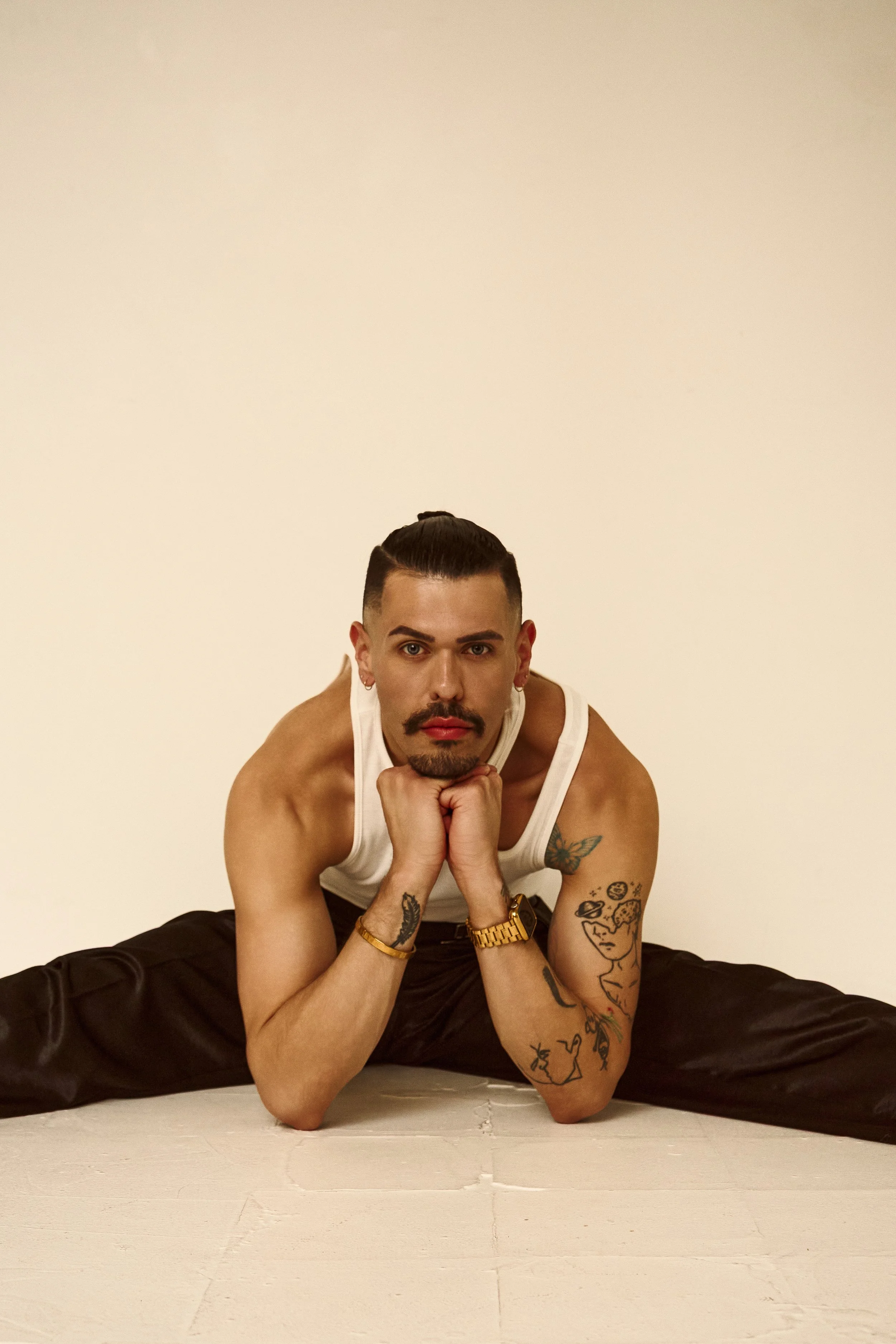 A young man with tattoos, a mustache, and earrings, wearing a white tank top and black pants, is sitting on the floor with legs spread wide, supporting his chin with his hands, in front of a plain beige background.