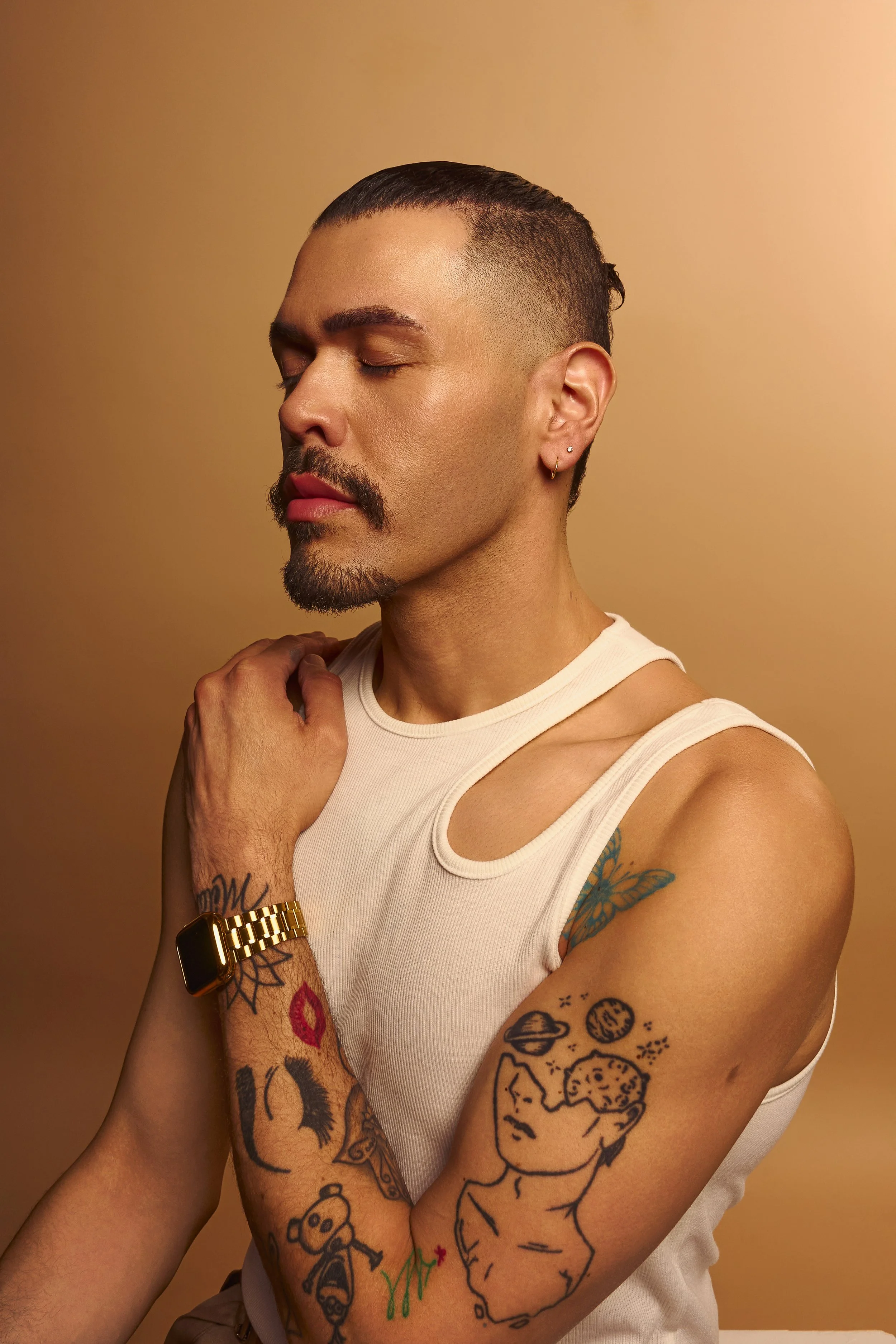 A person with a mustache and goatee, wearing a white sleeveless shirt and a gold watch, with tattoos on their arms and shoulder, closed eyes, and a neutral expression, standing against a gradient gold background.