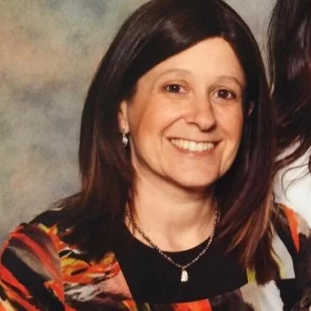 A smiling woman with short black hair in a blue top standing among green plants.