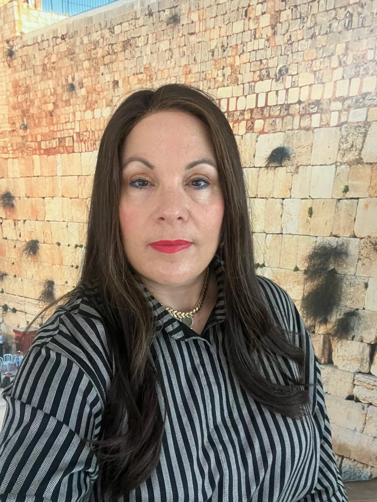 Woman with long dark hair wearing a black and white striped blouse and a gold chain necklace, standing in front of a large brick wall with some dark patches and mortar lines visible.