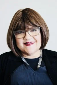 Portrait of a woman with short brown hair, glasses, and wearing a black top with a pearl necklace.