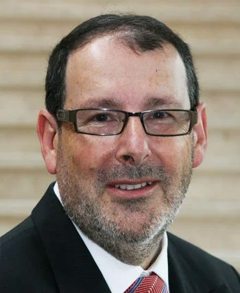A middle-aged man with short dark hair, glasses, and a beard, wearing a suit and tie, smiling against a wooden background.