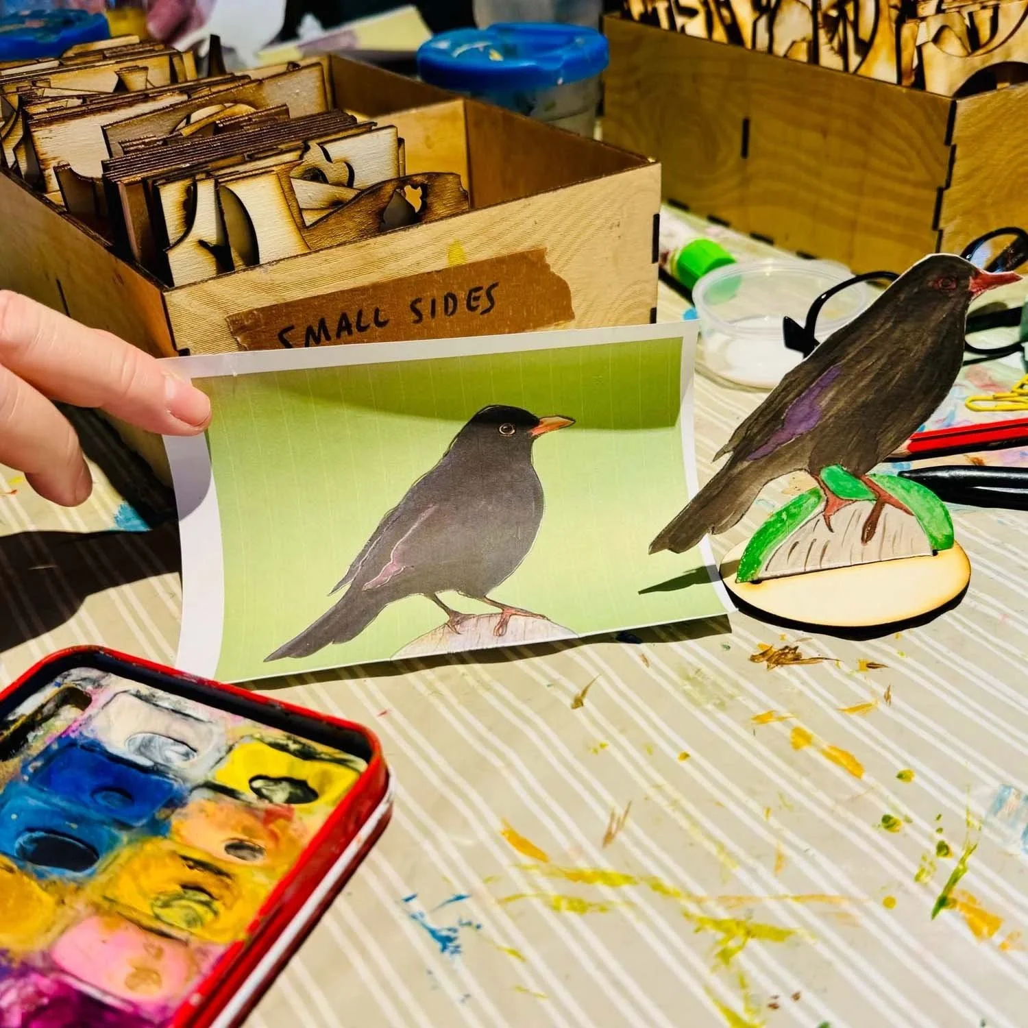 A person holding a printed illustration of a bird on a piece of paper. In front of the paper, there is a painted wooden bird figurine on a small base, both depicting a bird with dark feathers and an orange beak. The workspace is covered with a striped tablecloth and contains various art supplies, including a watercolor palette, brushes, and wooden boxes labeled 'Small Sides.'