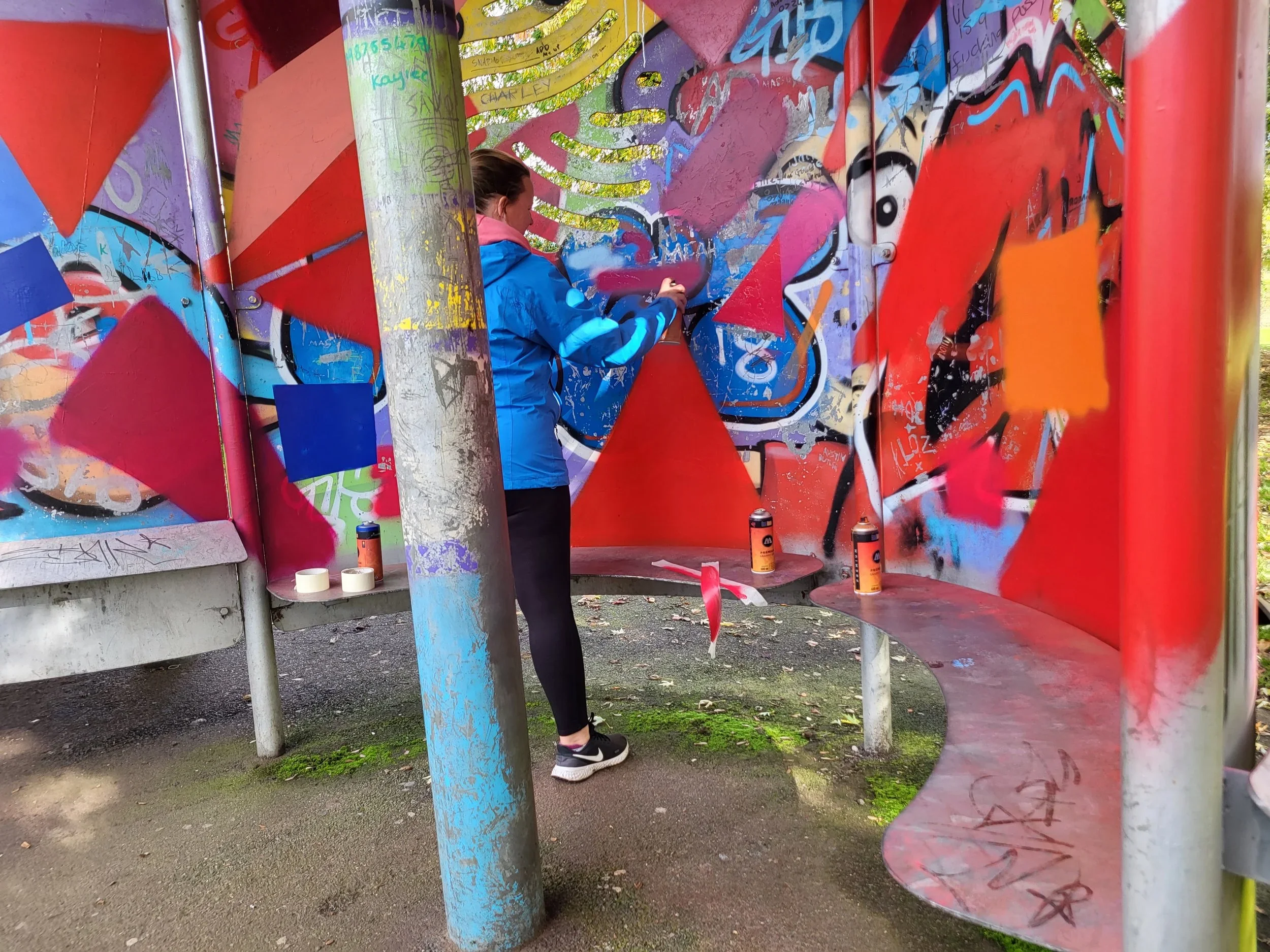 A woman in athletic clothing spray-paints graffiti on a colorful wall at an outdoor skate park.