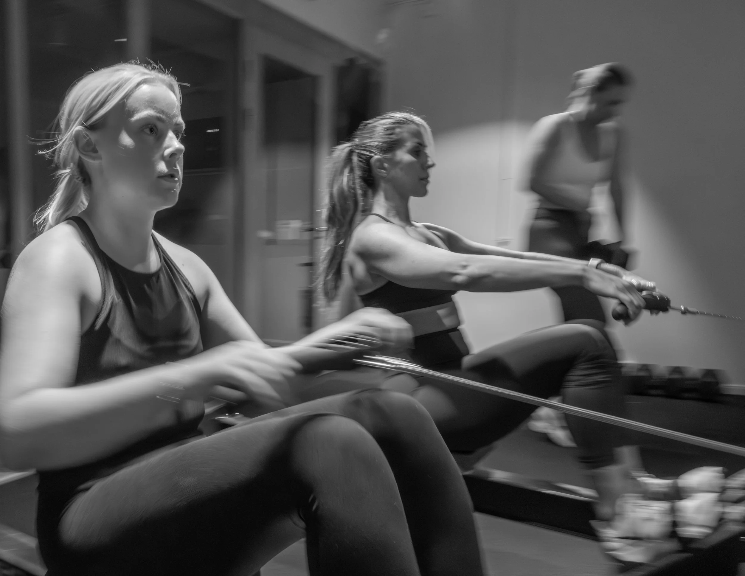Two women with athletic builds, wearing sleeveless tops, are engaging in an indoor fitness activity using rowing machines, with a trainer or observer standing behind them.