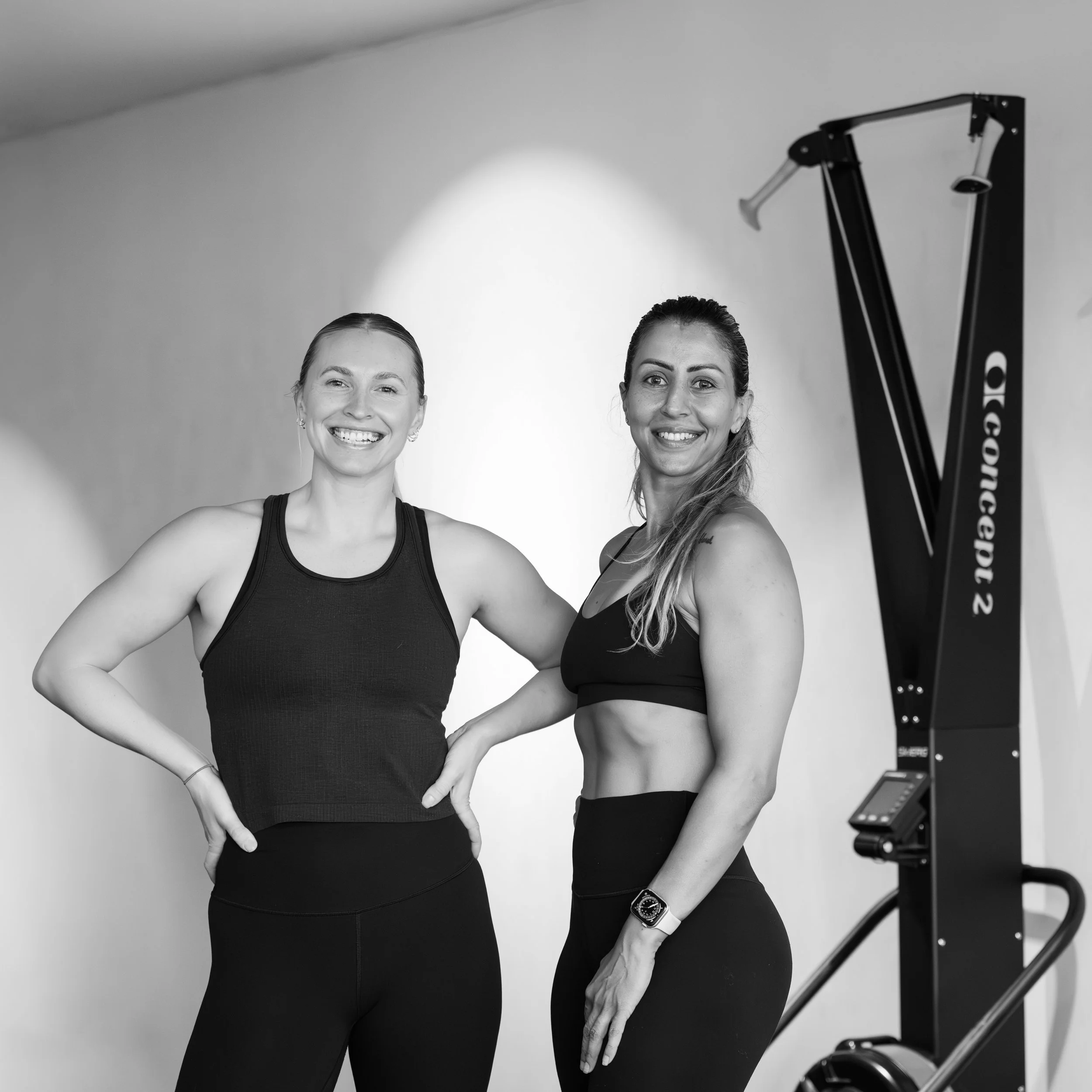Two women in athletic wear standing in a gym, smiling, with fitness equipment visible in the background.