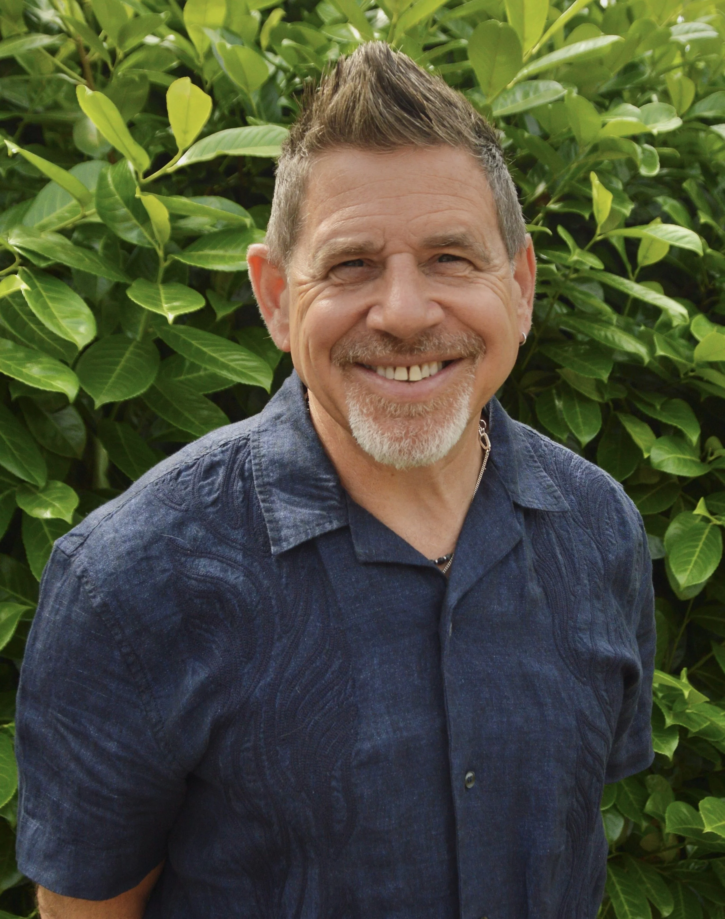 A middle-aged man with short gray hair, a goatee, and a mustache smiling outdoors in front of lush green foliage. He is wearing a dark blue, textured, button-up shirt.