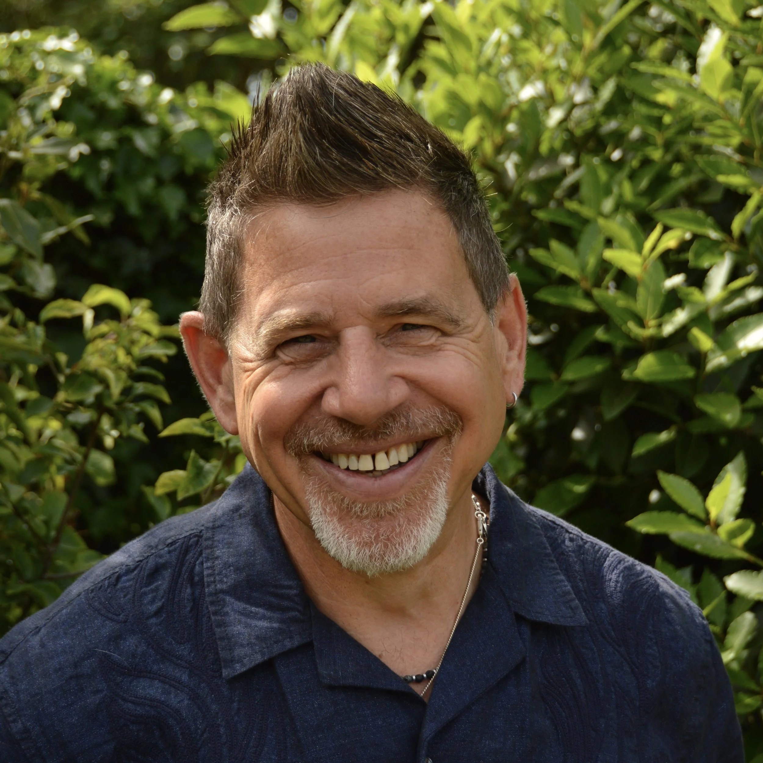 Close-up of a smiling man with gray beard, wearing a dark blue textured shirt and a silver necklace, standing outdoors in front of green leafy bushes.