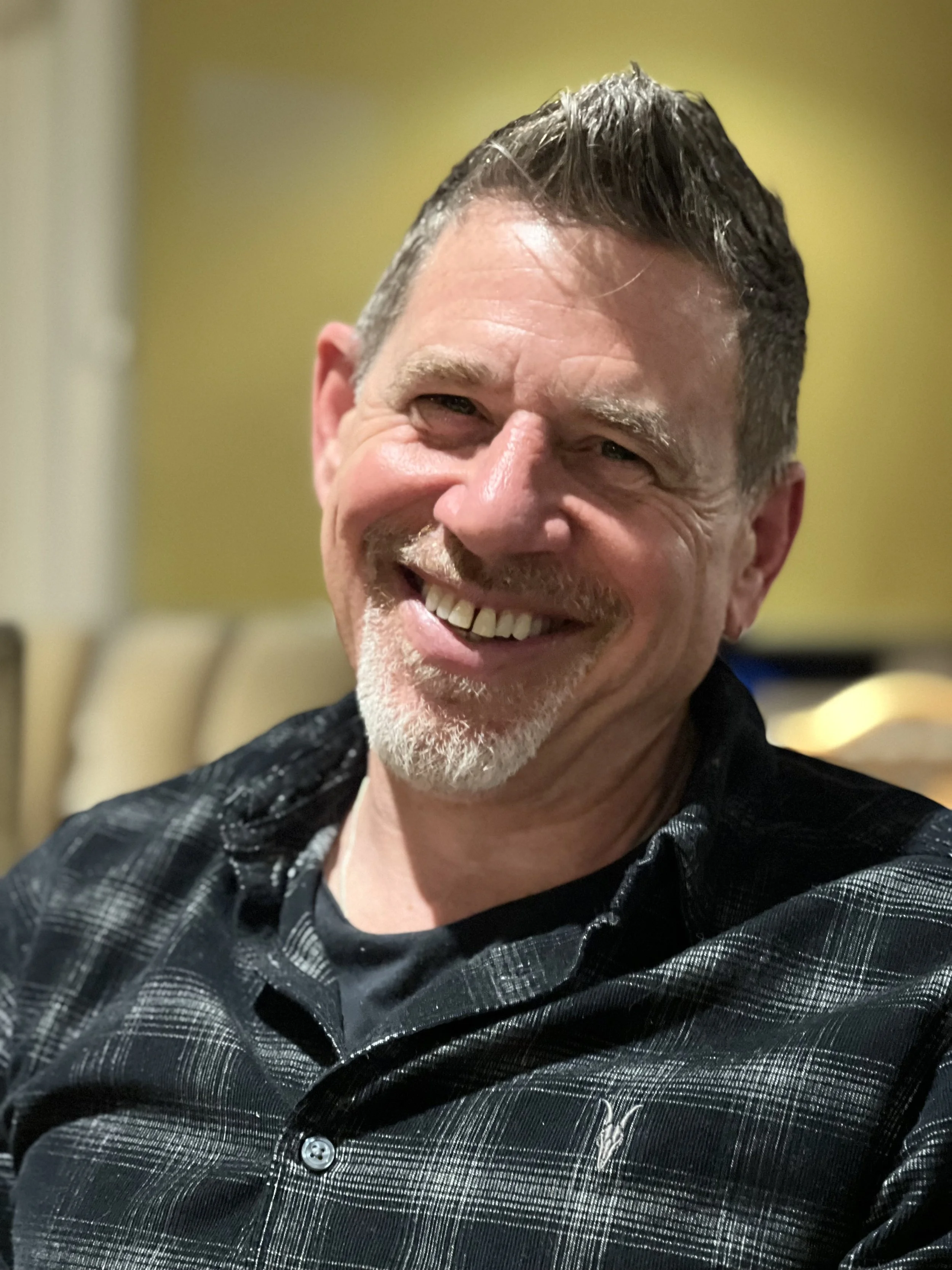 A smiling middle-aged man with short, gray hair and a beard, wearing a black and gray checkered shirt.