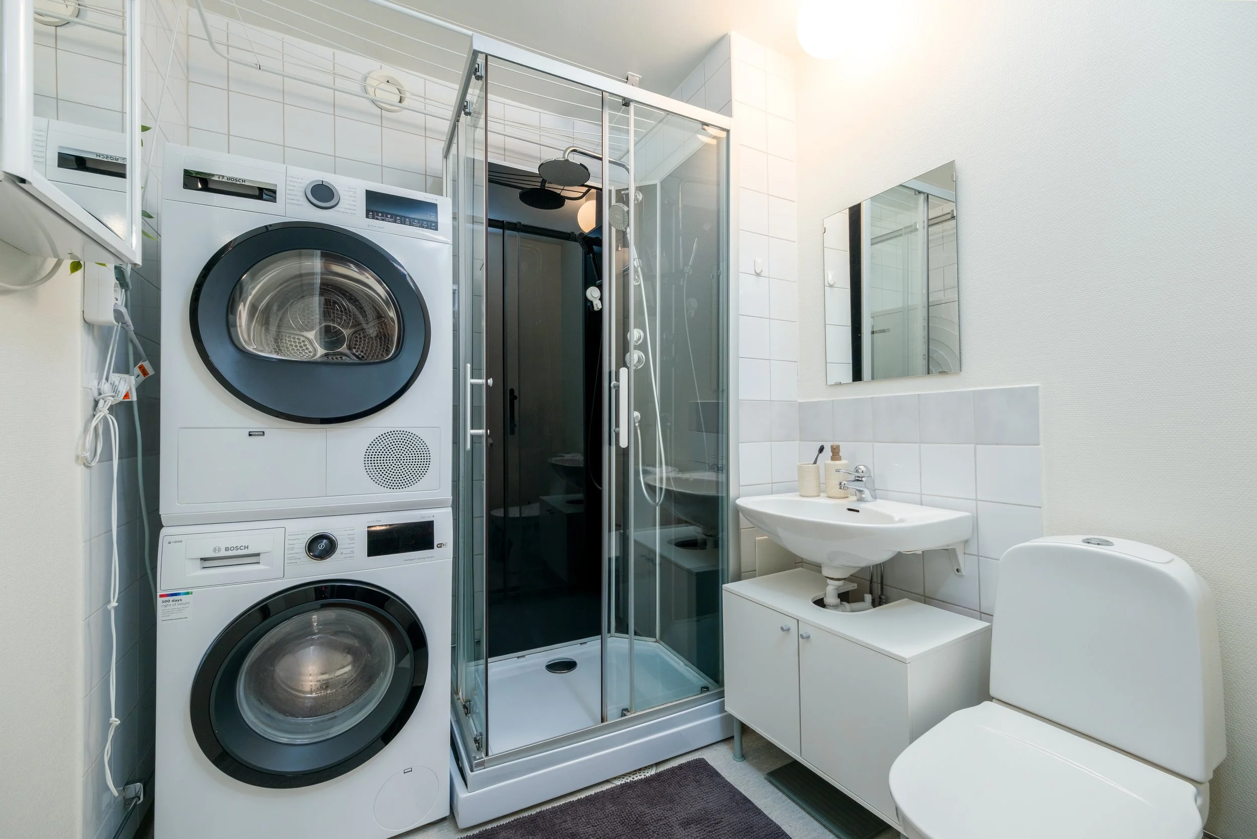 Compact bathroom with stacked washing machine and dryer, shower with glass doors, small sink with a mirror above, and a toilet.