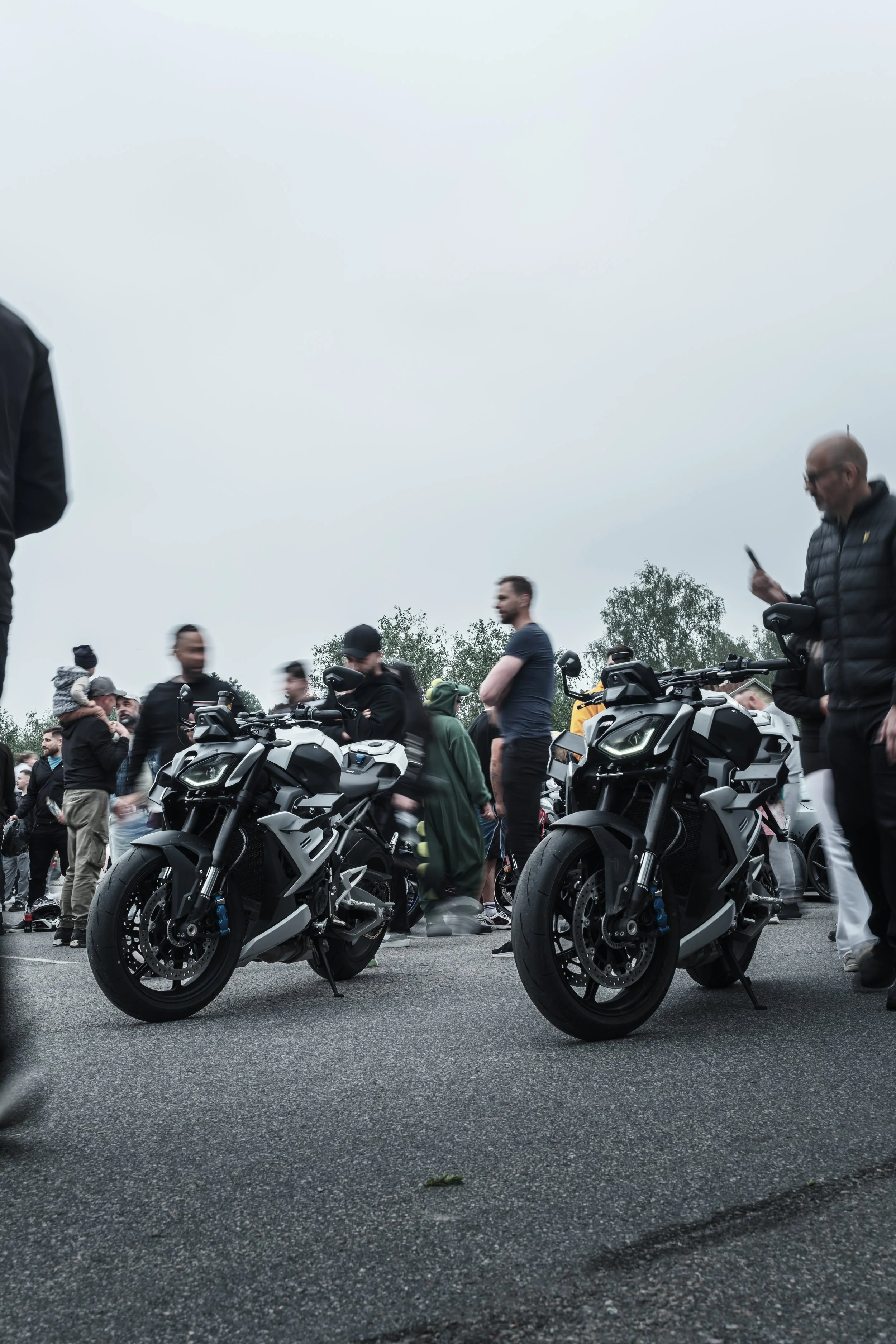Two electric motorcycles parked on a wet road surrounded by a crowd of people at an outdoor event on a cloudy day.