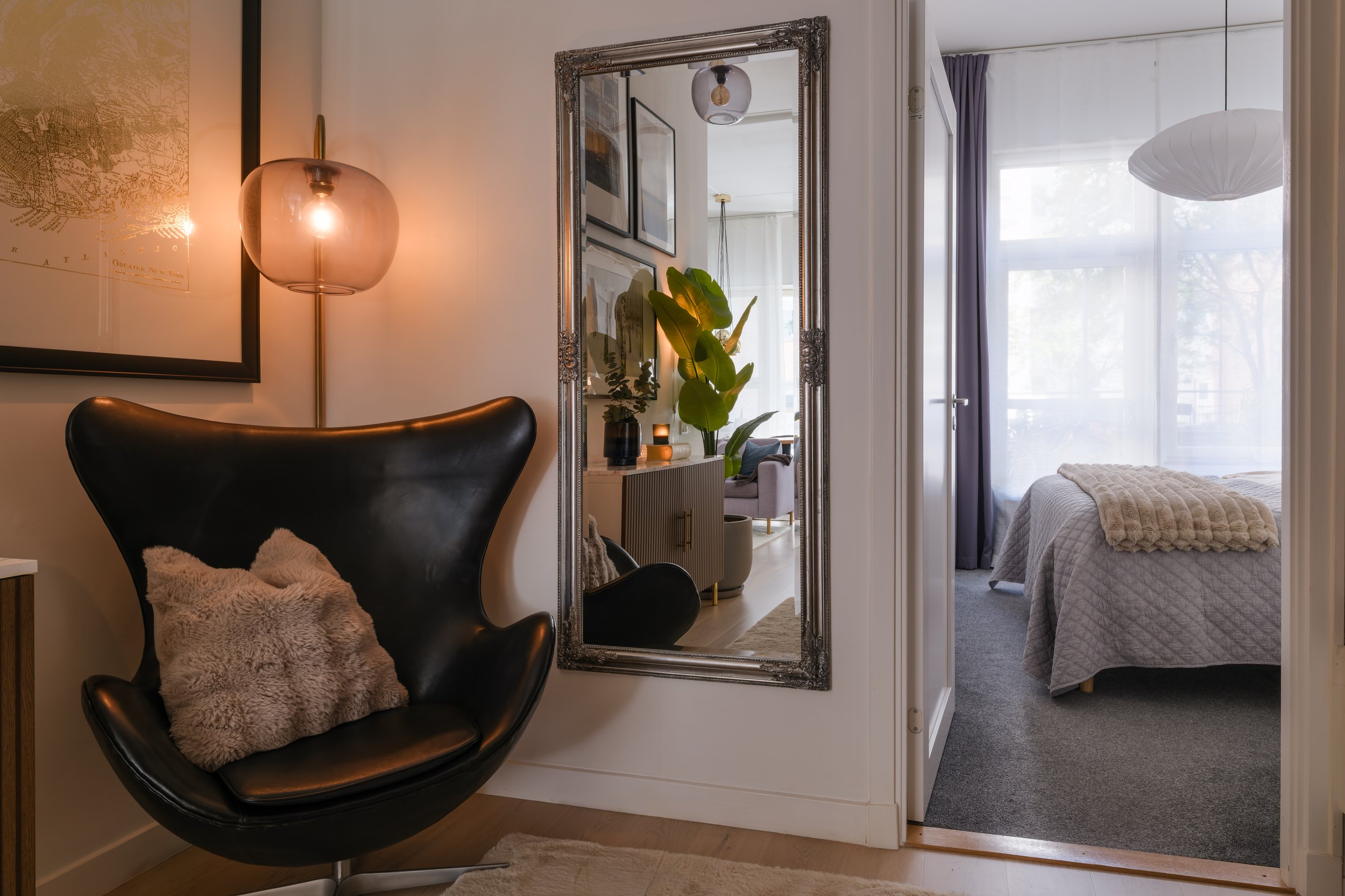 Interior detail photograph of a black leather egg chair beside an ornate mirror, warm lamplight, view through to the bedroom.