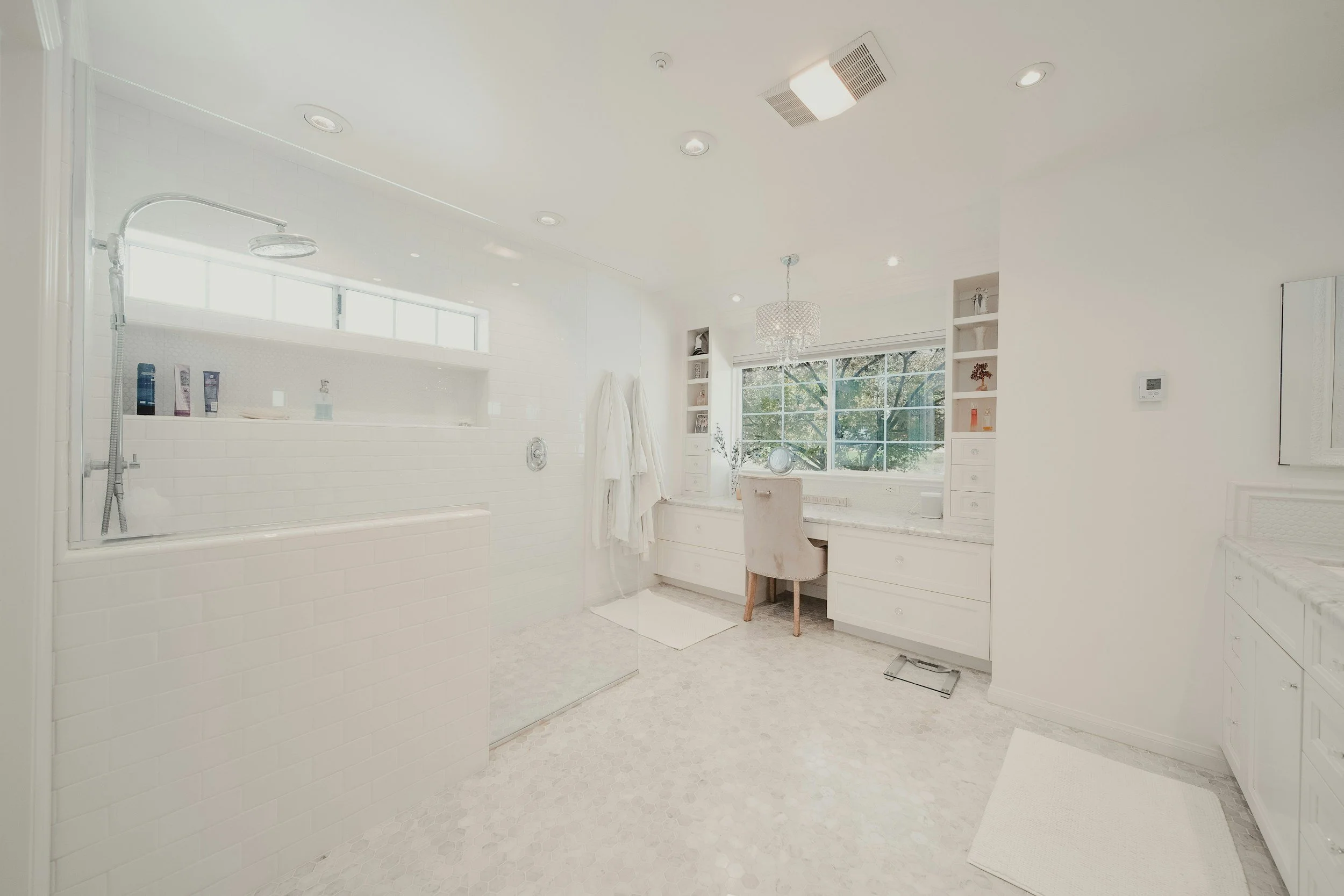 Bright, white bathroom with a walk-in shower, a vanity desk with storage drawers, and a large window that overlooks trees.
