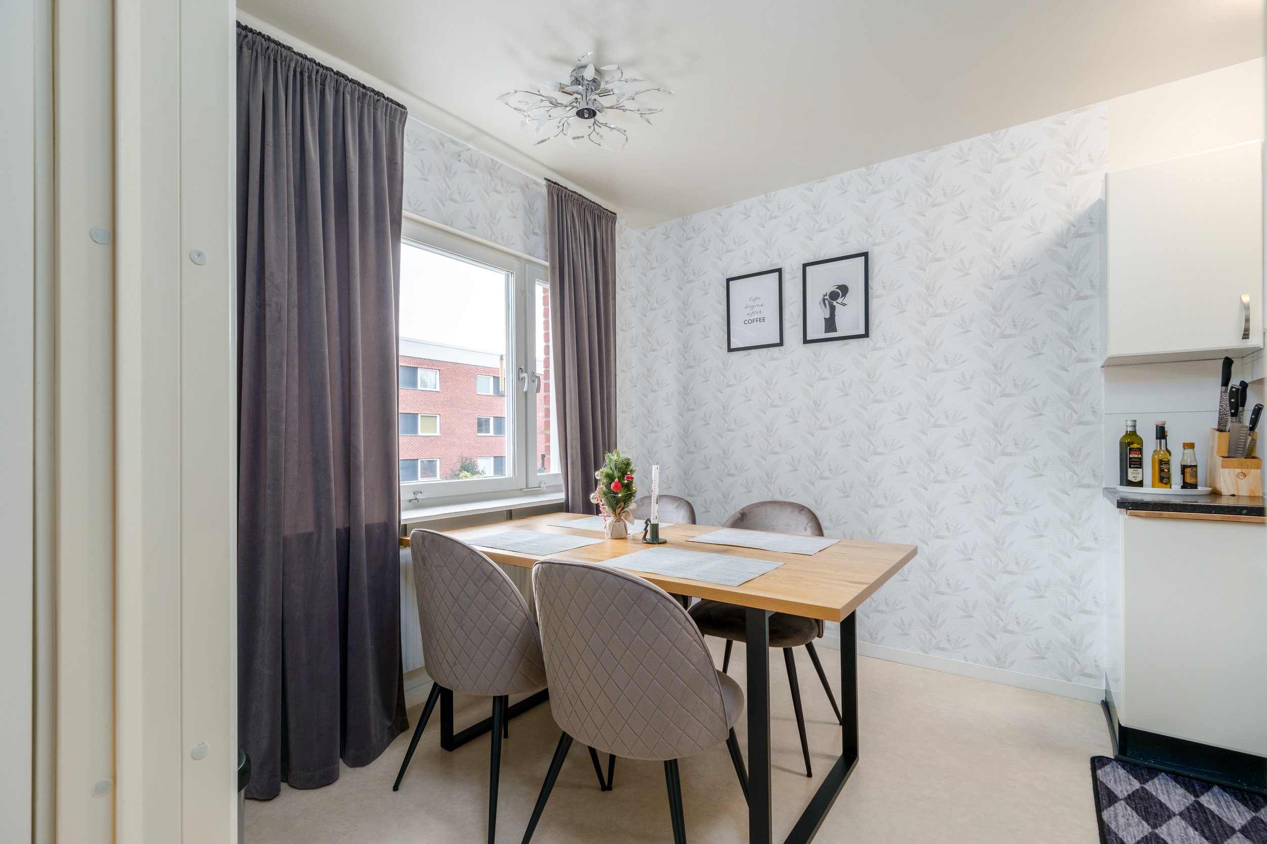 Dining room with wooden table, four beige chairs, window with gray curtains, wallpaper with leaf pattern, wall art, and a small Christmas tree centerpiece.
