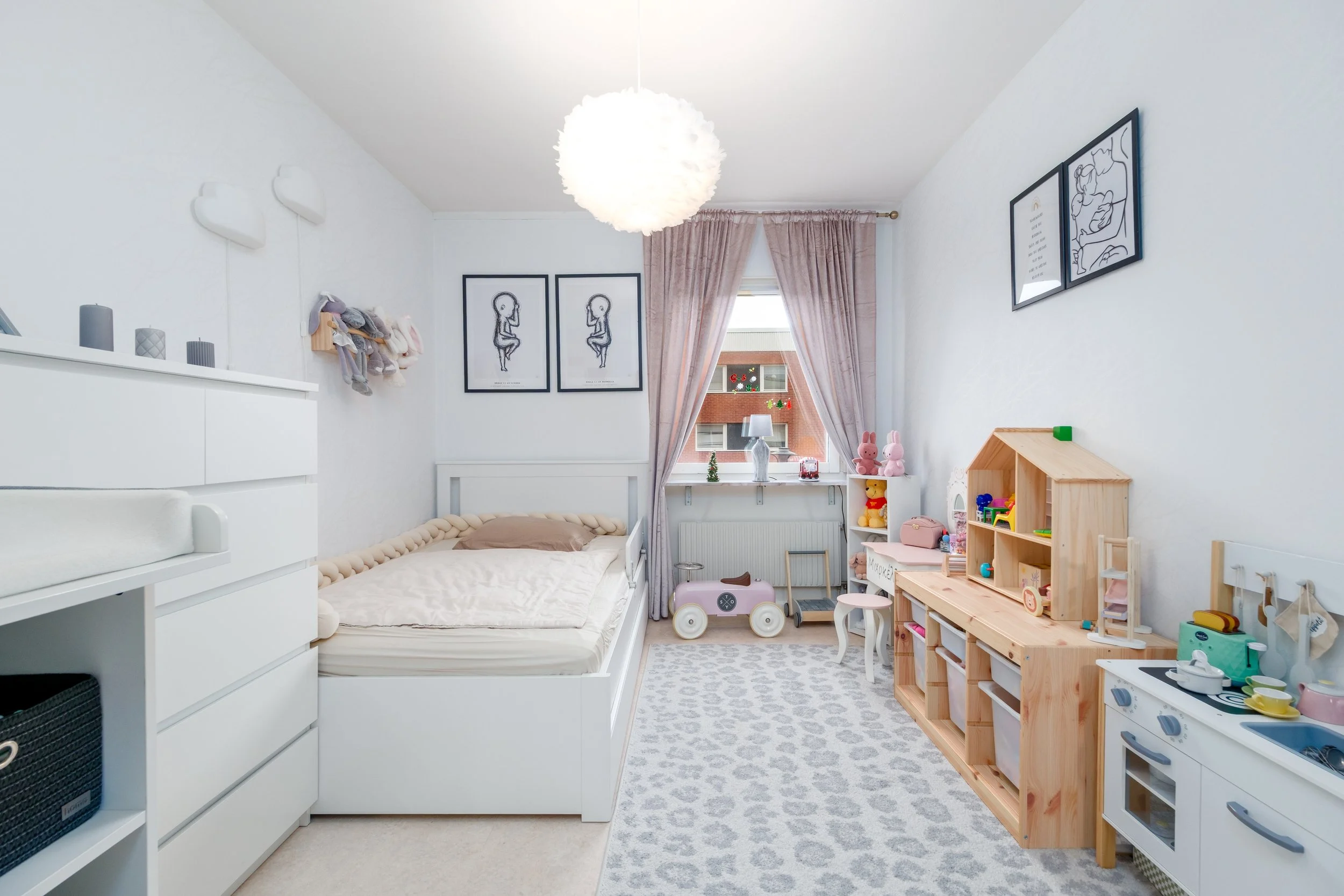 Child's bedroom with white walls, a bed, stuffed animals, a play kitchen, and toys, decorated with minimalistic black and white artwork.
