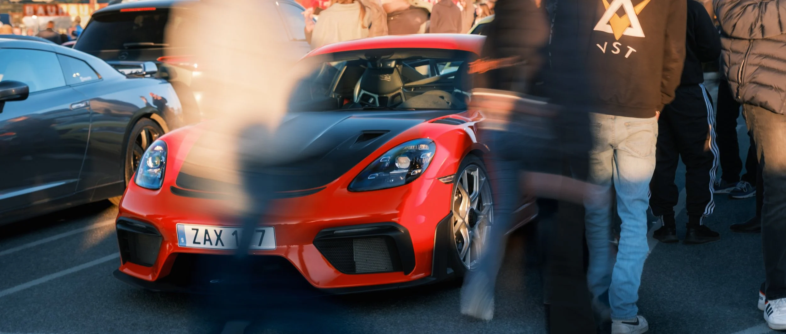 Red and black sports car parked among other vehicles with people standing around at an outdoor event.