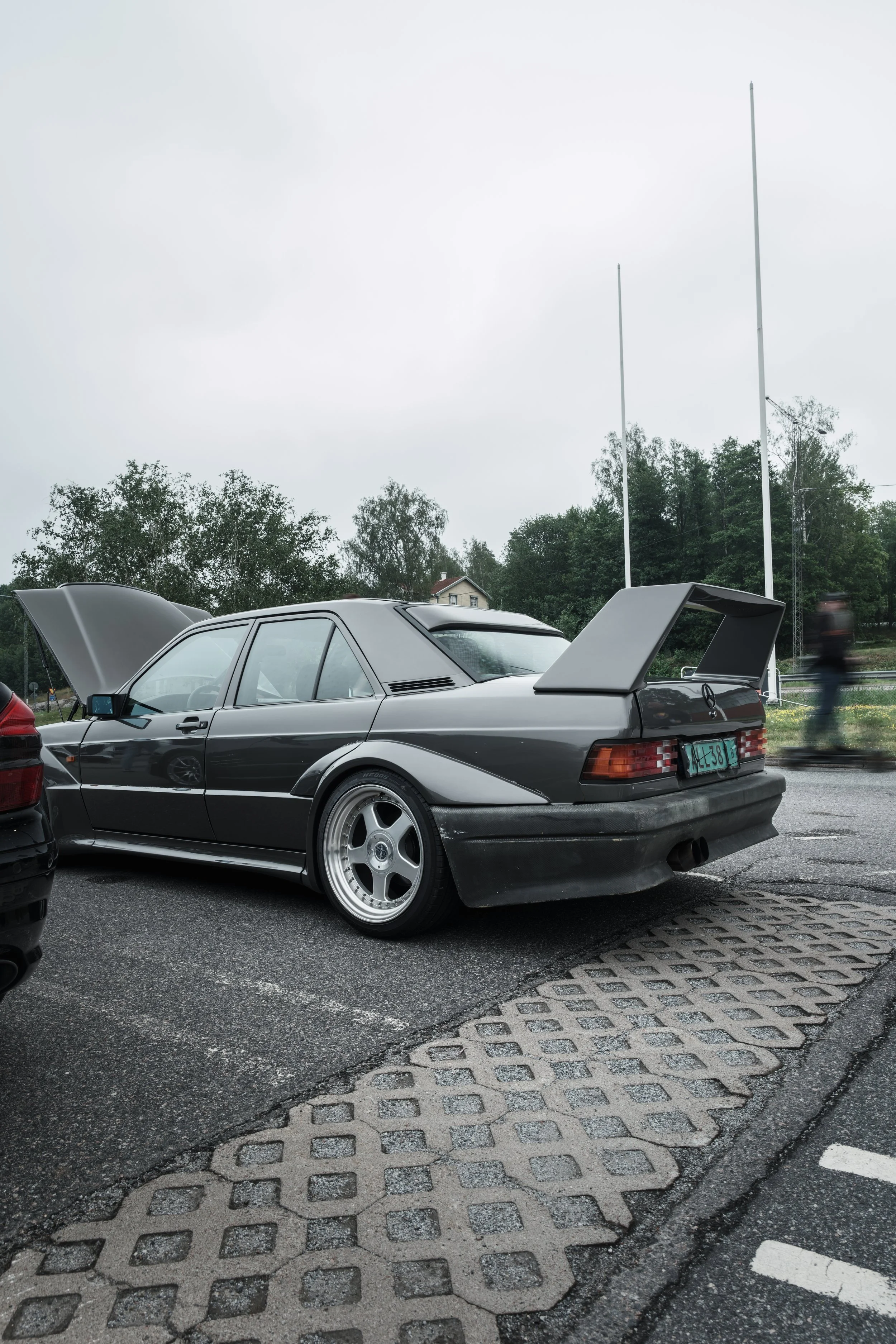A vintage black sports car with the hood and rear hatch open, parked on a parking lot on an overcast day, with trees and a person blurred in the background.