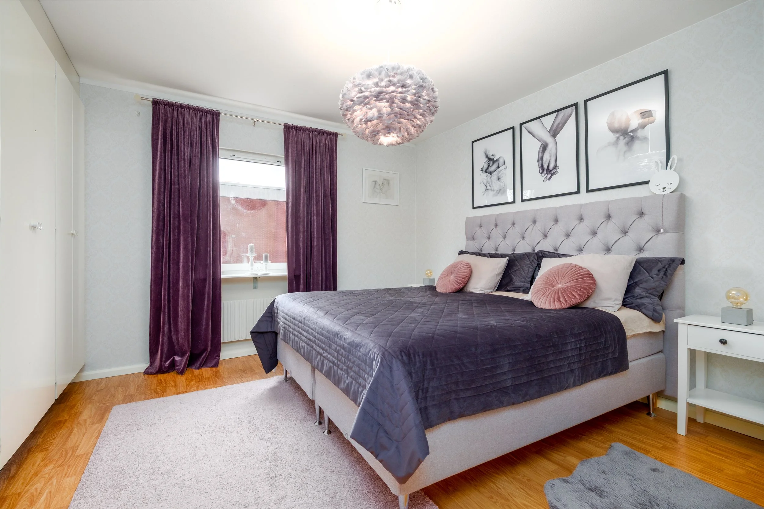A modern bedroom with a large upholstered bed, purple curtains, wall art, and a pink feathered ceiling light fixture.