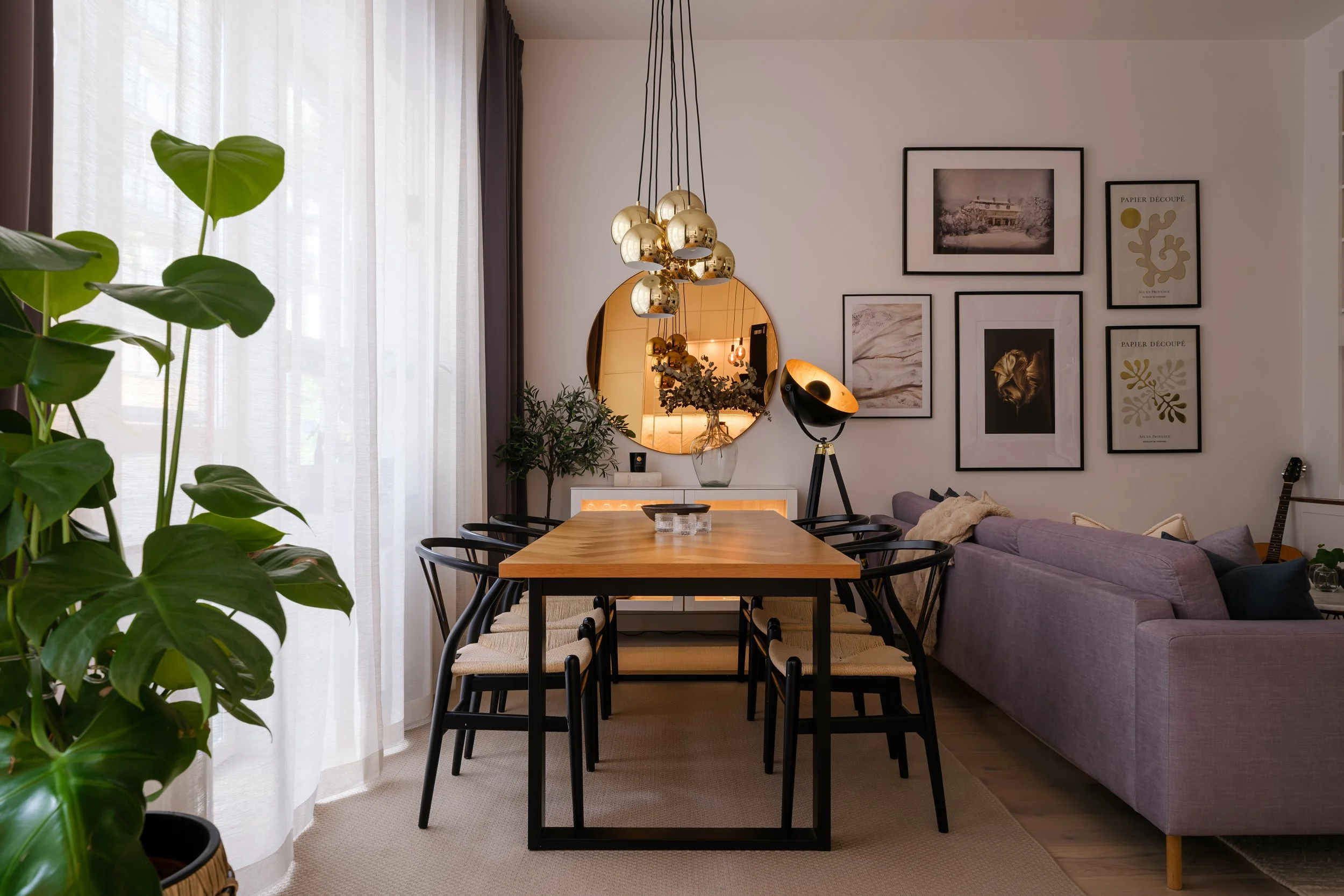 Editorial interior photograph of a dining area with round gold mirror, tripod lamp and gallery wall, warm evening styling — Stockholm apartment.