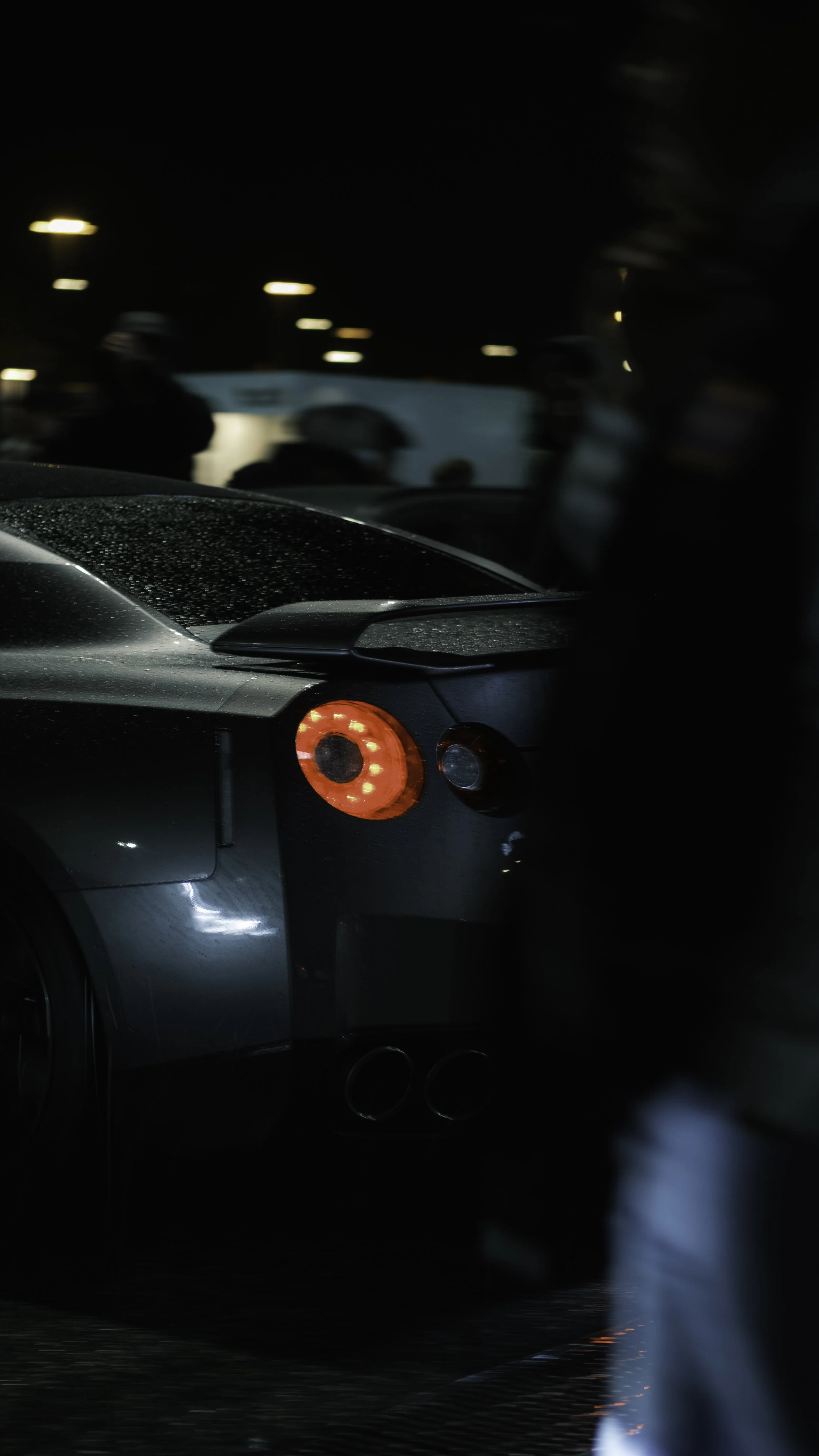Close-up of the rear of a black sports car with illuminated red taillights at night, with blurred background lights and wet road surface.
