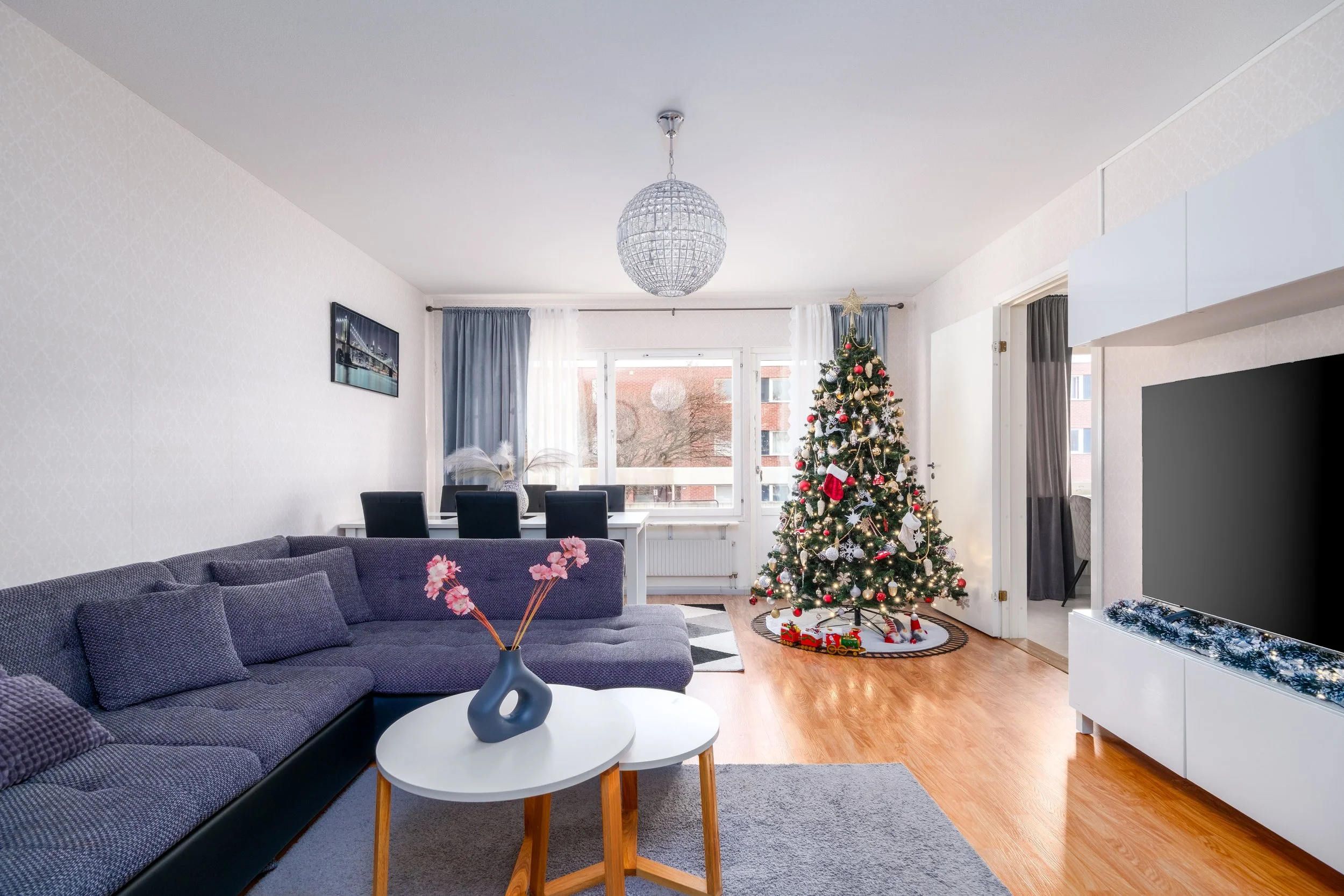 Living room with a Christmas tree decorated with ornaments, presents underneath, a large flat-screen TV, a dark gray sofa with pillows, small white and wood tables, a vase with pink flowers, a dining area, with a window and balcony door, decorated with blue curtains, a chandelier, and a gray area rug.