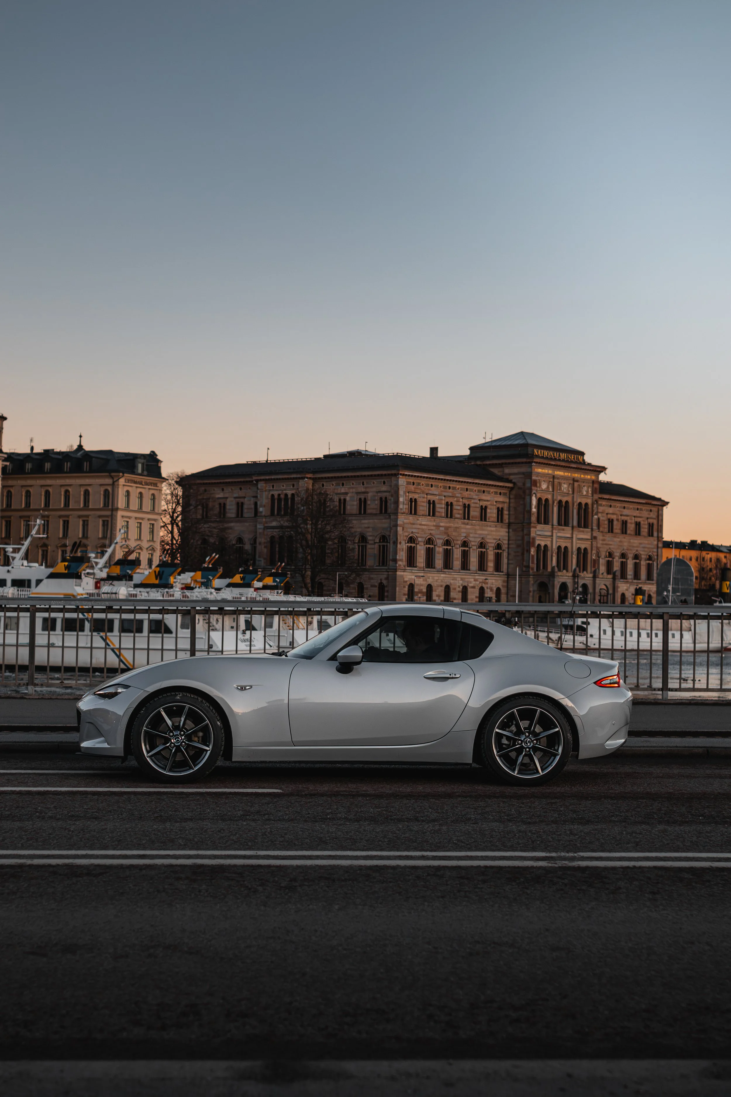A silver convertible sports car parked on a city street in front of a river and historic buildings at sunset.