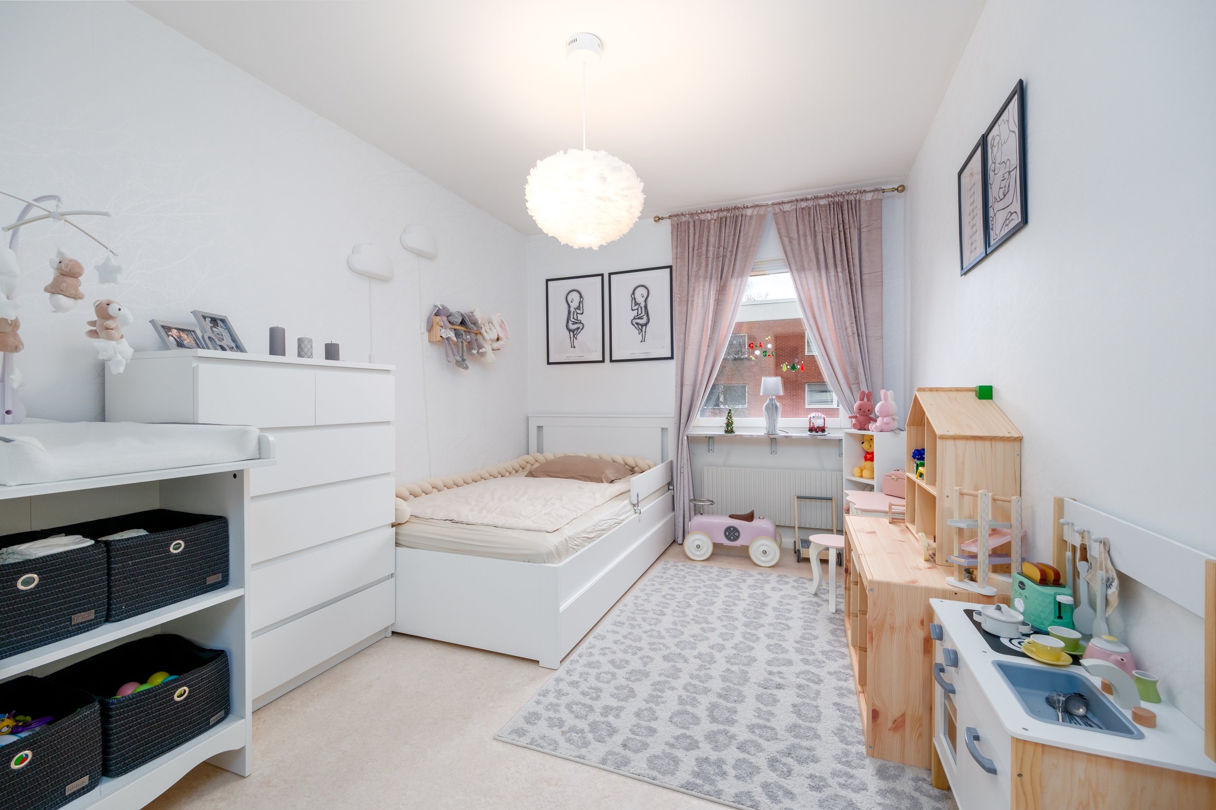 A child's bedroom with white walls and furniture, pink curtains, toys including stuffed animals and a toy kitchen, framed artwork, a light fixture, and a window with a view of a building outside.