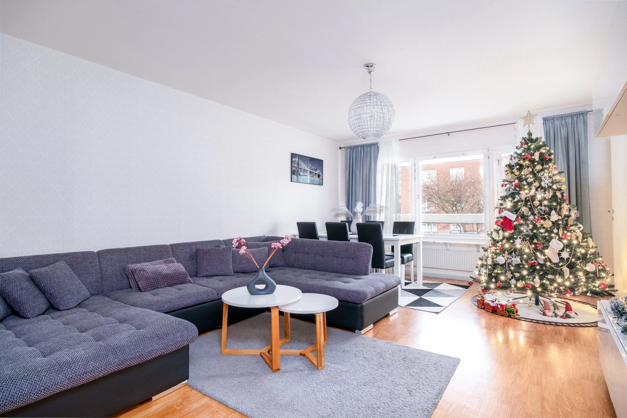 Living room decorated for Christmas with a decorated Christmas tree, white and gold ornaments, stockings, and lights. There is a large L-shaped gray sofa, a small round white coffee table with a decorative vase and flowers, a dining table with black chairs, and a window with blue curtains.