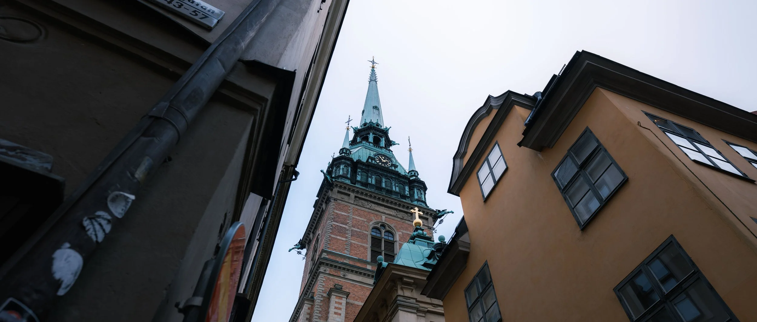 Looking up between tall buildings at a clock tower with a spire and crosses, showing a cloudy sky in the background.