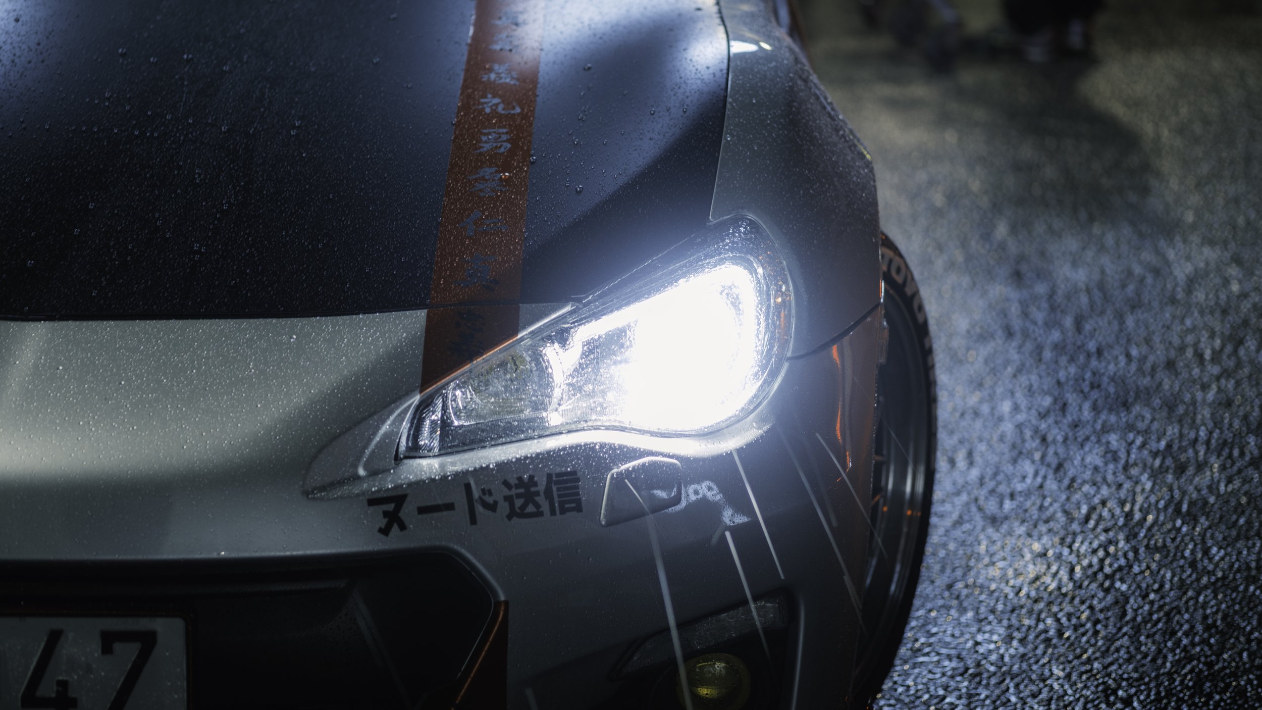 Close-up of a silver sports car's front with a bright headlight on, wet road surface, reflections, and Japanese text decals on the car