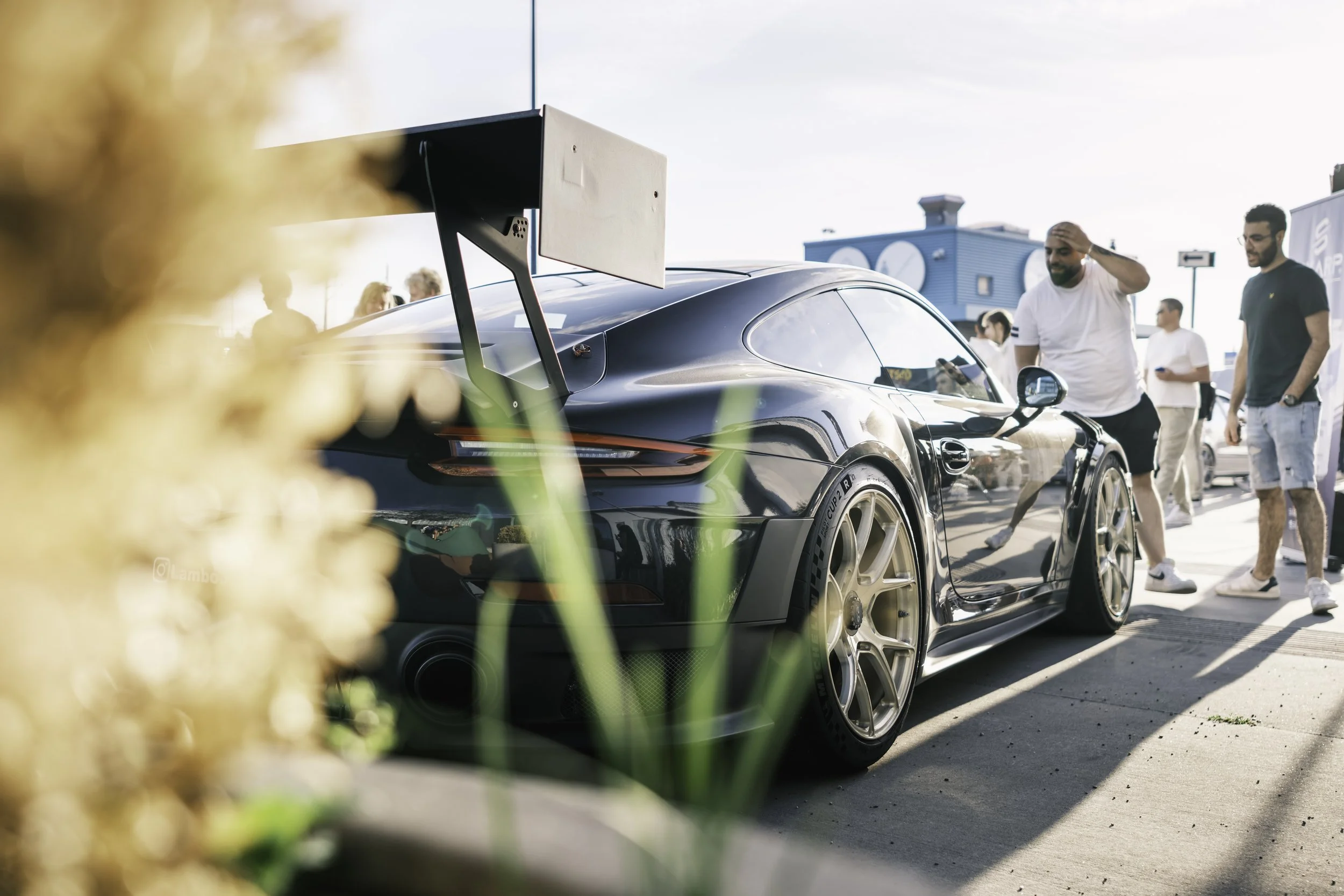 A black race car with a large rear wing on display at a car show, surrounded by people, with a blue building in the background and some plants in the foreground.