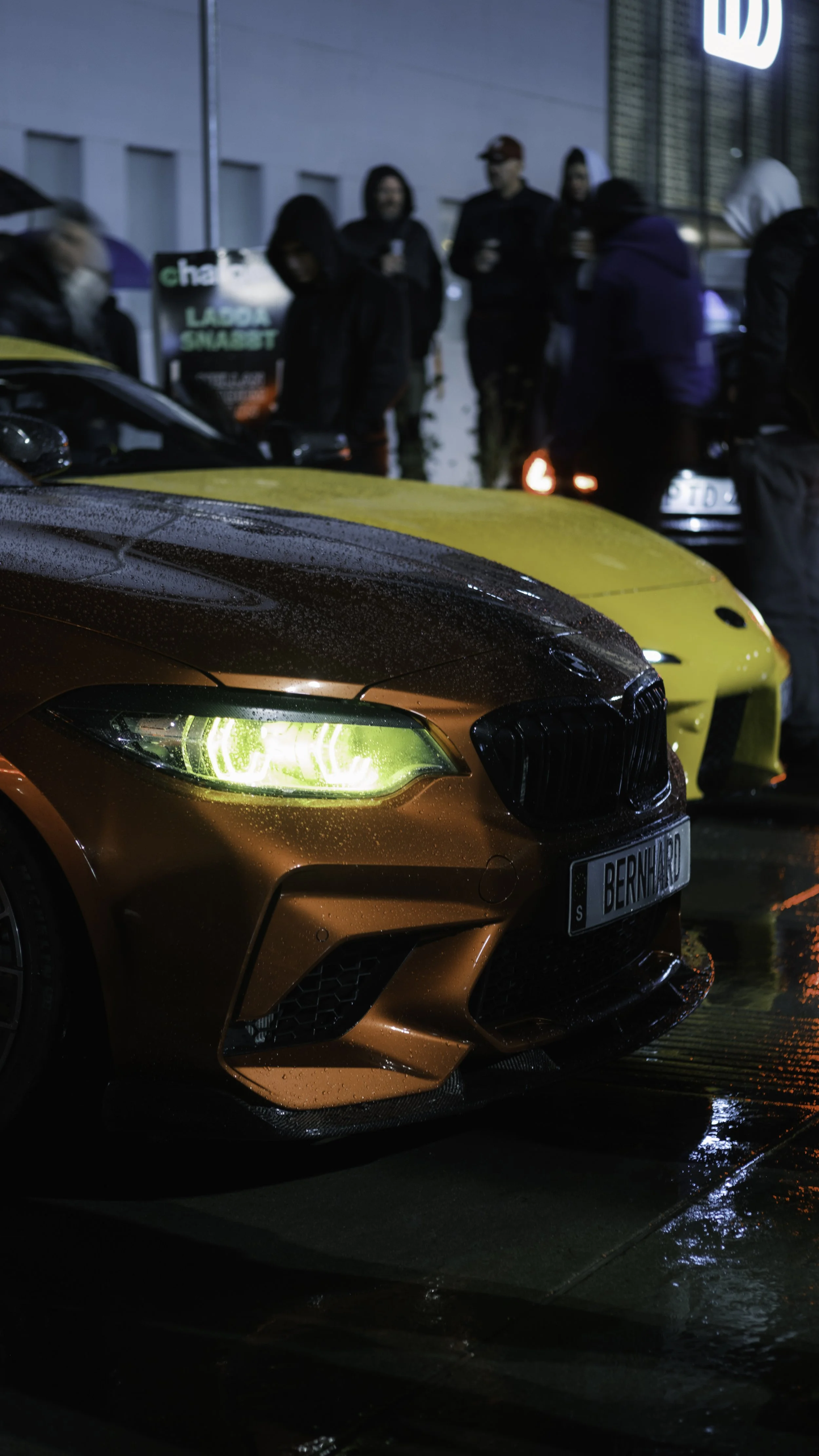 A group of people gather around two luxury sports cars parked outside on a rainy night. The foreground features a metallic bronze and black BMW M4 with blurred rain droplets on the hood and tire, illuminated by nearby lights. The background shows several individuals, some holding drinks, standing near a yellow car and a black car, with a building and a lit sign behind them.
