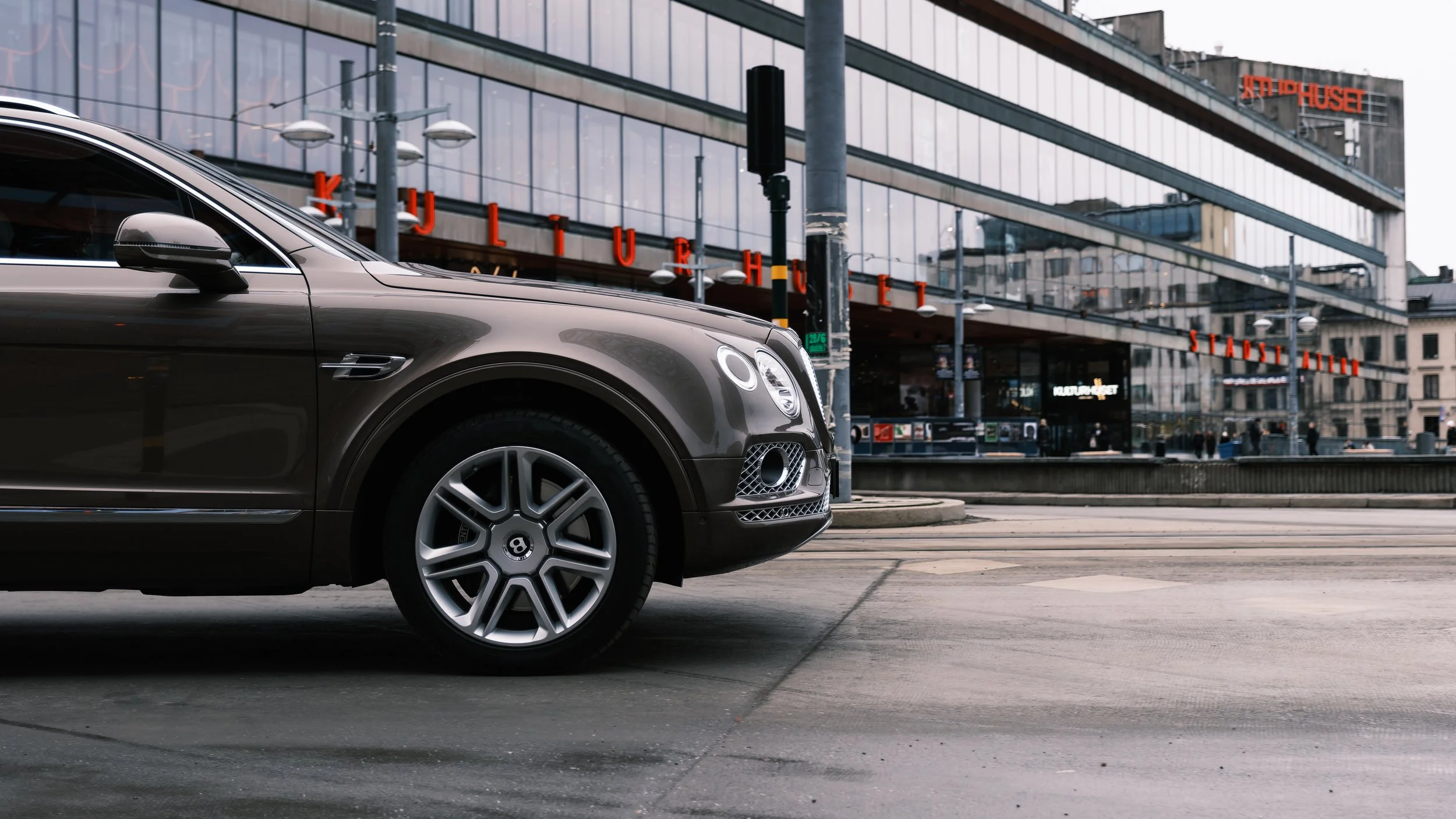 Part of a luxury car in front of building with red signs and glass facade in an urban setting.